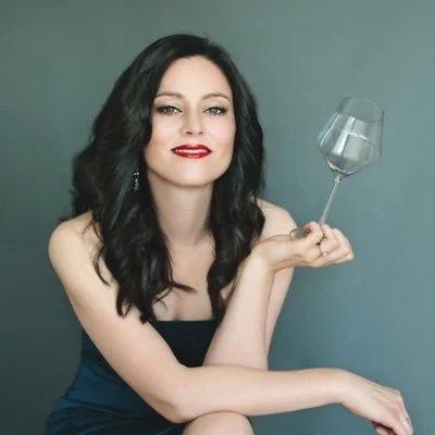 A woman with long dark hair, wearing red lipstick and a black dress, sitting with her arm resting on her knee, holding an empty wine glass in one hand against a plain gray background.