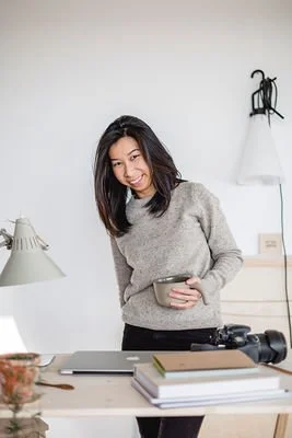 Woman standing at a desk holding a bowl in a room with white walls, a lamp, a white dress on a hanger, and a camera on the desk.