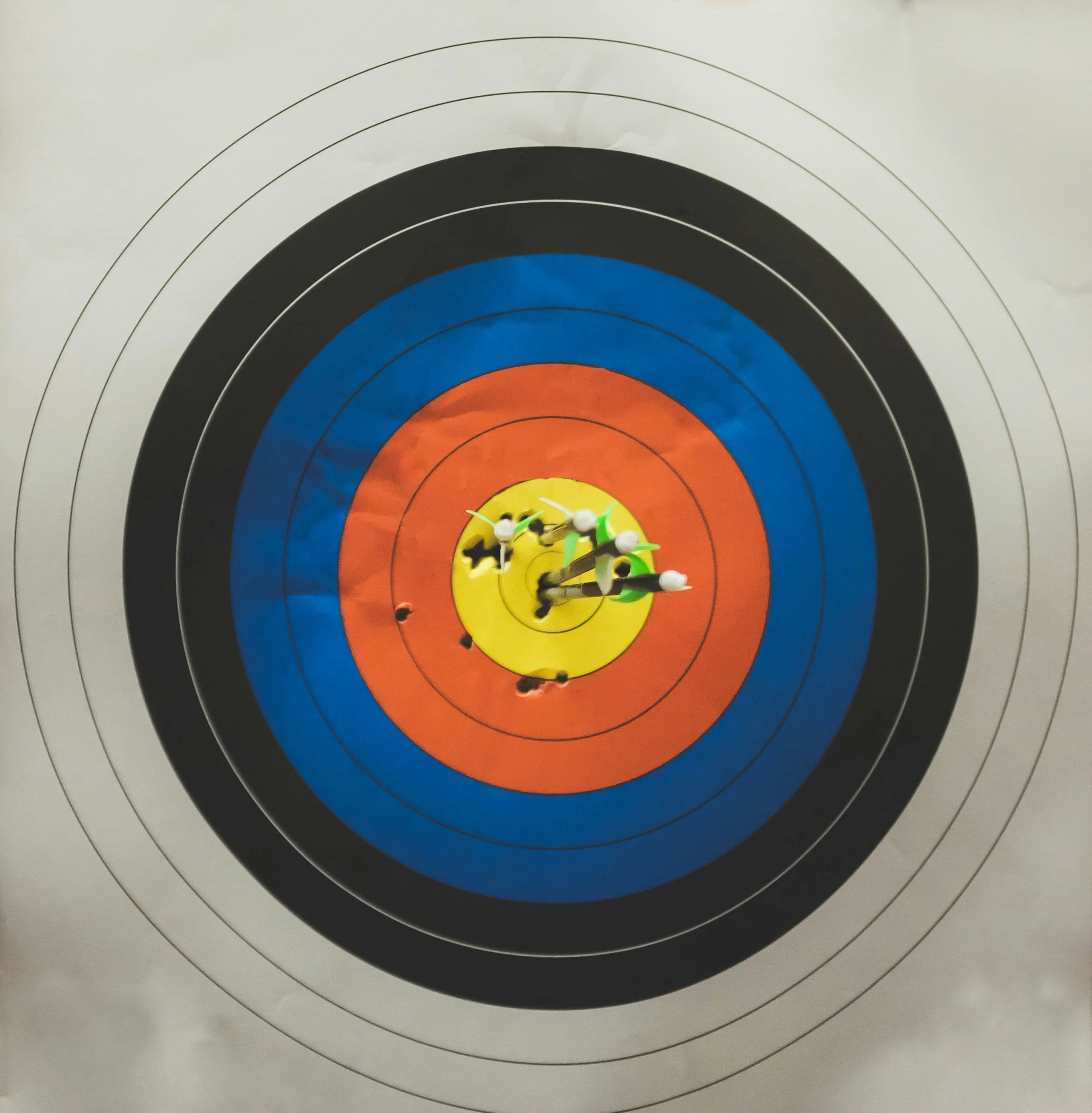 An archery target with colored rings and a clock in the center. The target has multiple arrows stuck in the yellow and orange rings.