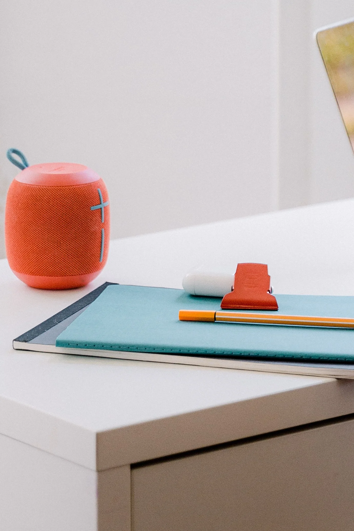 A white desk with a pink portable speaker, several notebooks, a pen, a white tube, and a red clip, with part of a laptop or tablet visible in the upper right corner.