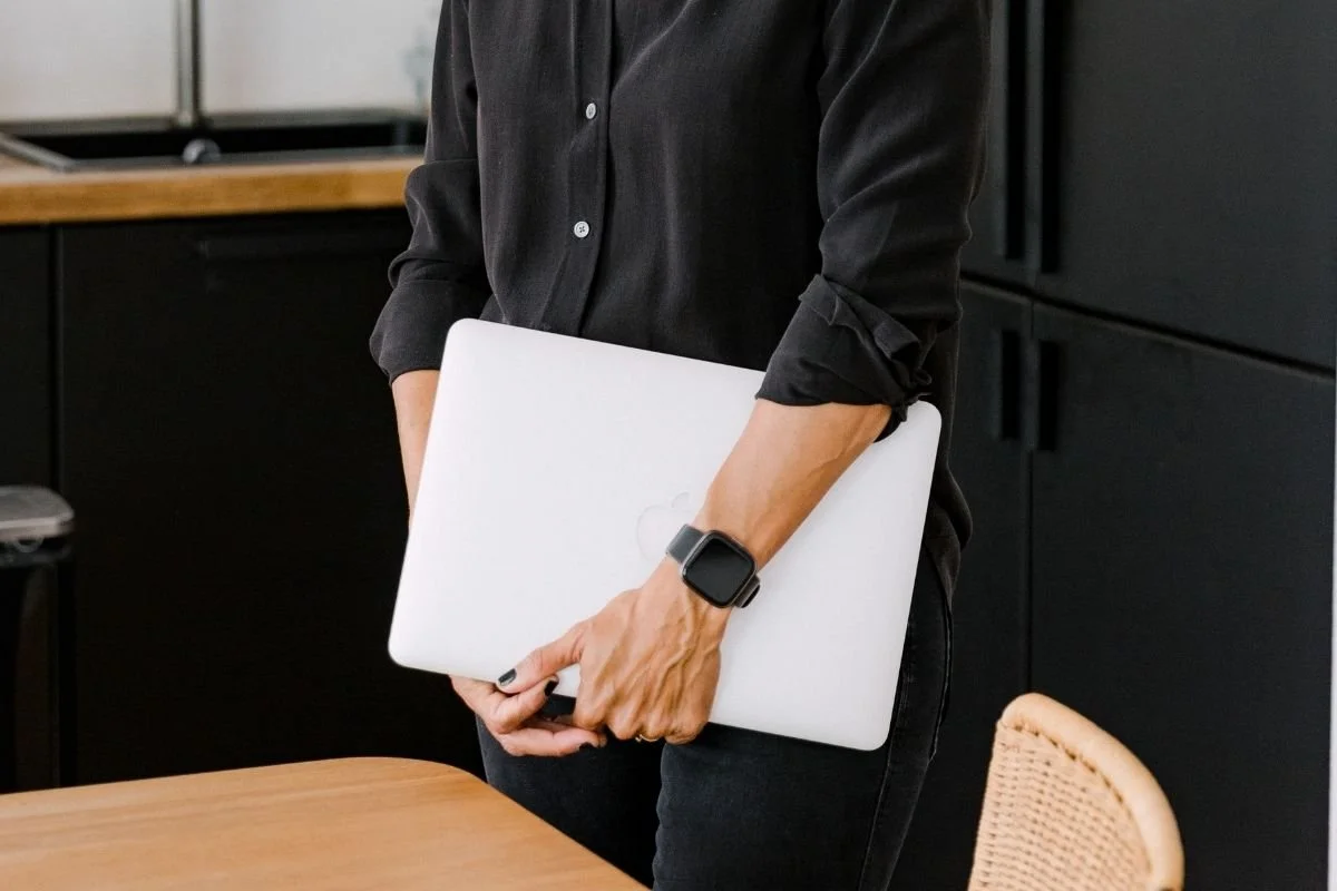 Person in black shirt holding a closed silver laptop and wearing a smartwatch, standing in a modern office kitchen area.