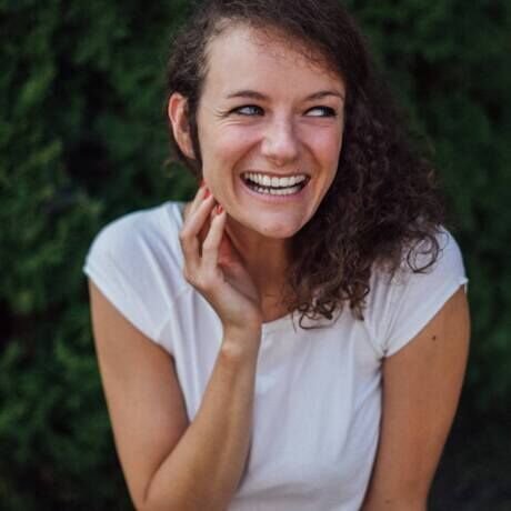 A woman with curly brown hair smiling and looking to the side, wearing a white shirt, outdoors with greenery in the background.