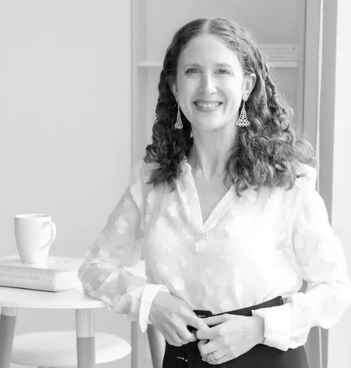A woman with curly hair wearing a white blouse and chandelier earrings, smiling while standing next to a small table with a coffee mug and a book in a bright room.