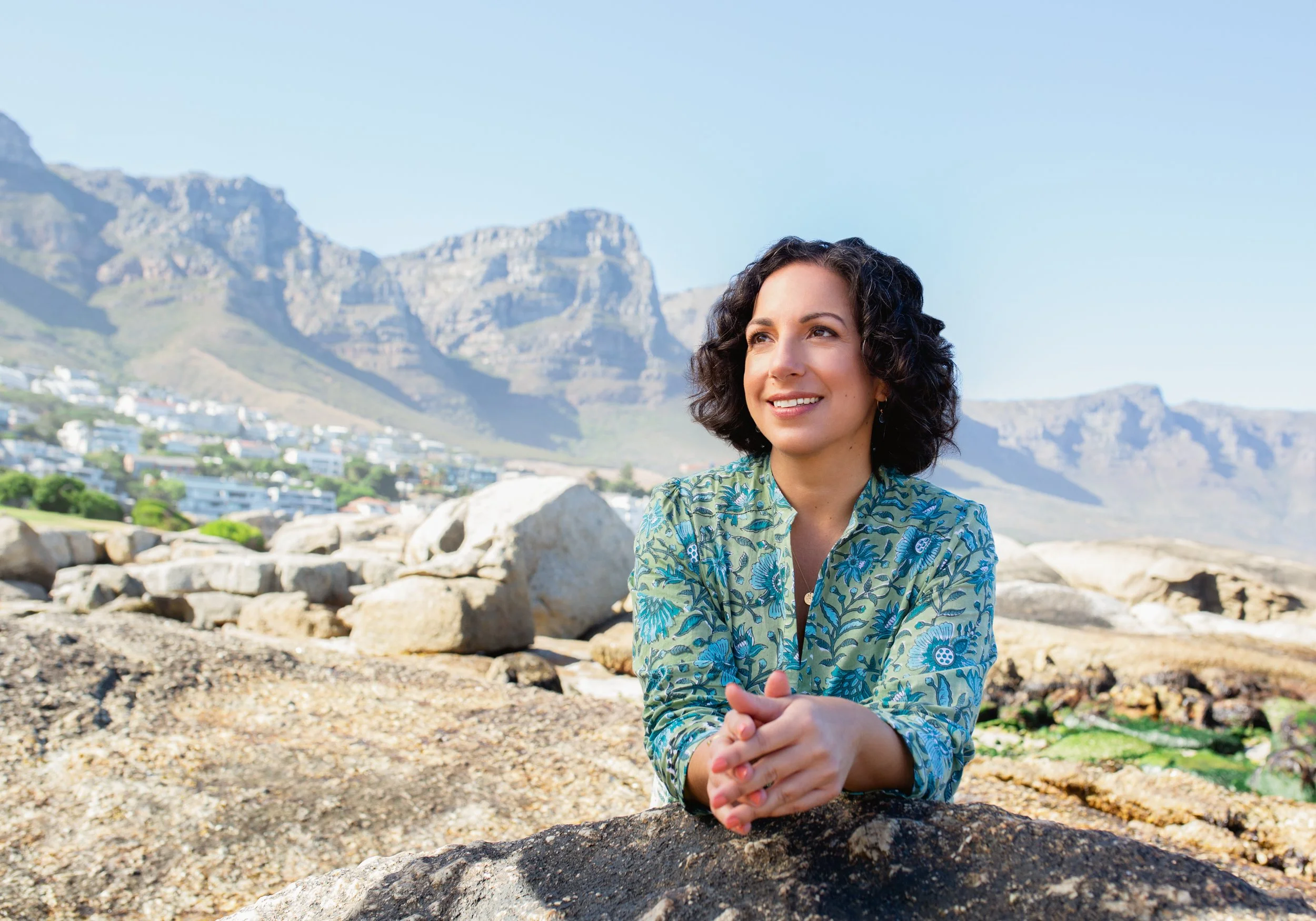 A woman with curly dark hair sitting on rocks by the ocean with mountains and a town in the background, wearing a green floral blouse, smiling and looking to her left.