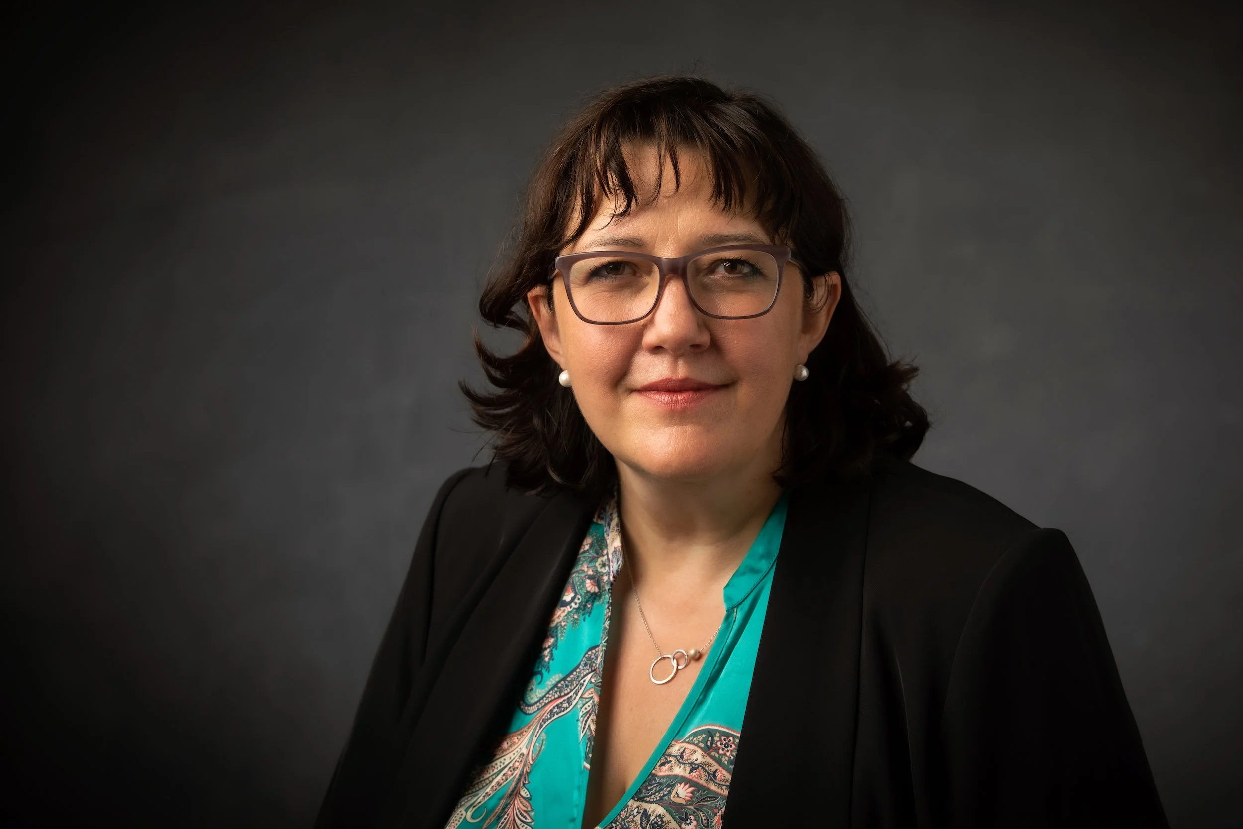 A woman with dark brown hair, glasses, and pearl earrings wearing a black blazer over a turquoise patterned blouse, posing against a dark gray background.