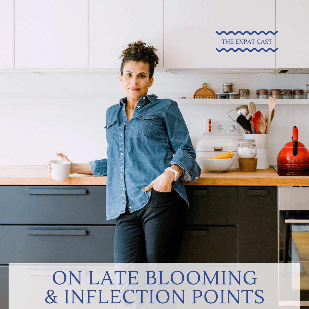 A woman with short curly hair in a denim shirt and black pants, standing in a modern kitchen holding a white mug, with a serious expression. The kitchen has white cabinets, a wooden countertop, and various utensils and jars. There's a text overlay that reads 'On Late Blooming & Infection Points' and a label 'The Expat Cast' in the top right corner.