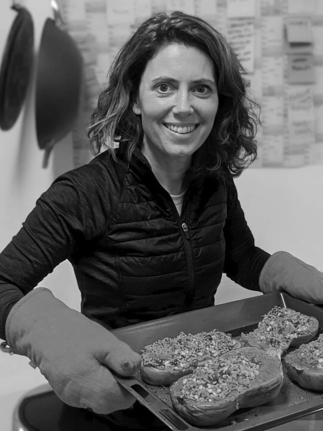 A woman smiling and wearing oven mitts, holding a baking tray with bread or pastries topped with nuts or seeds, in a kitchen setting.