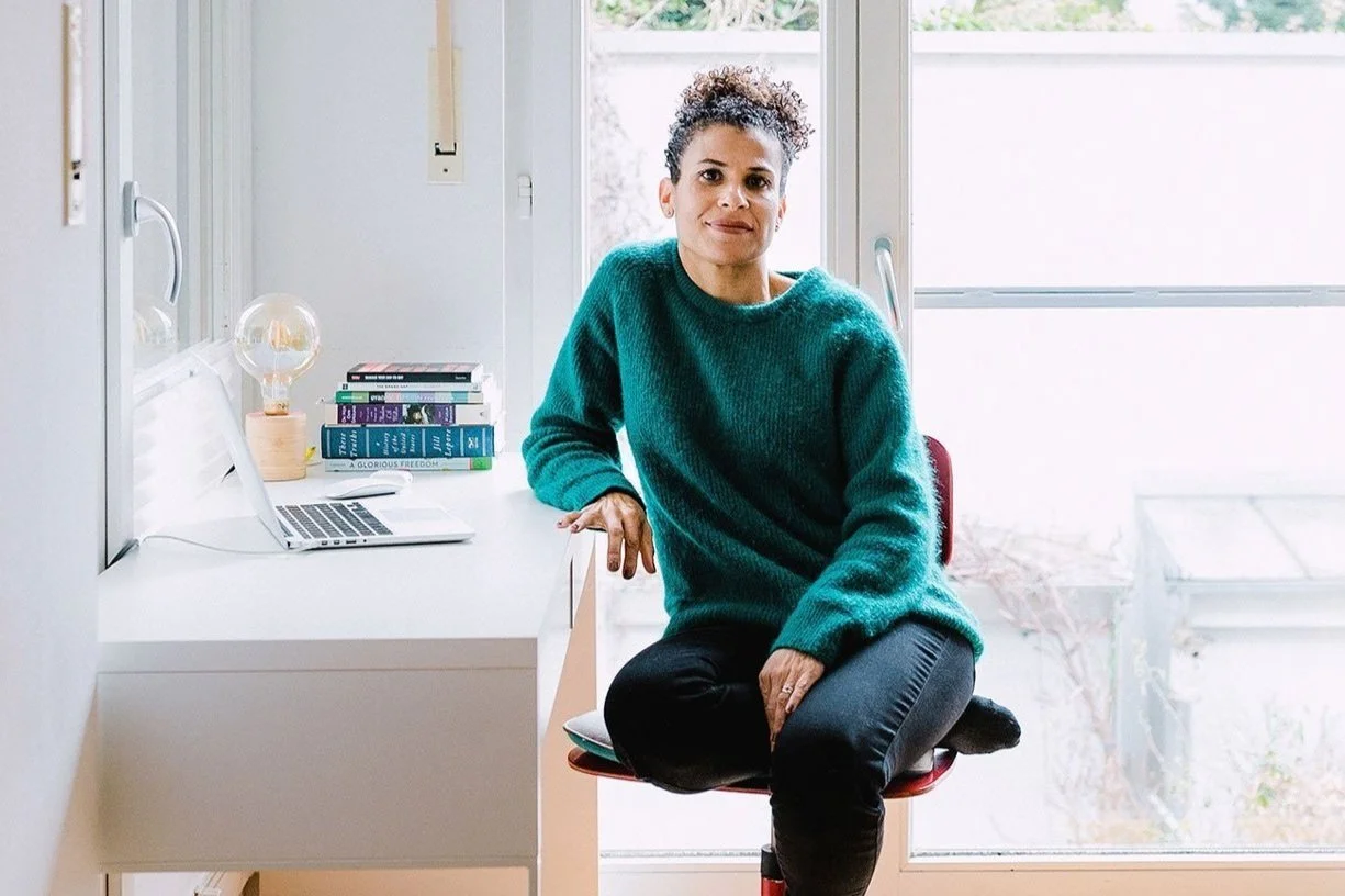 Woman sitting on a red chair at a white desk with a laptop, a stack of books, a lamp, and a laptop charger inside a room with large windows.