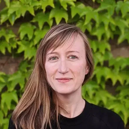 A woman with long, light brown hair standing in front of green foliage, wearing a black top and smiling gently.