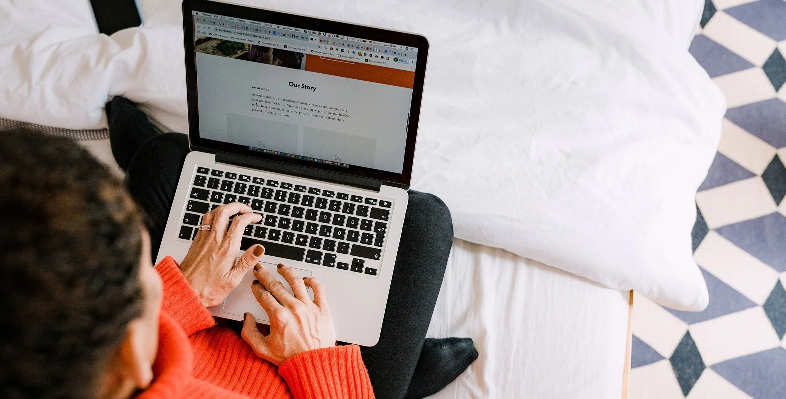 A person in a red sweater using a silver laptop on their lap, sitting on a bed with white bedding and geometric patterned blue and white rugs.
