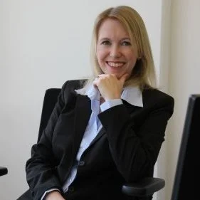 A smiling woman with blonde hair wearing a black blazer and white shirt, sitting at a desk in an office setting.