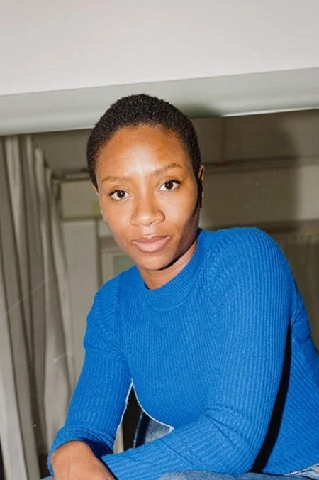 A woman with short curly hair wearing a blue knit sweater, looking at the camera indoors.
