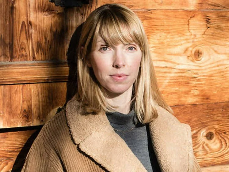 A woman with shoulder-length blonde hair and bangs, wearing a tan coat and a dark shirt, standing indoors against a wooden wall.