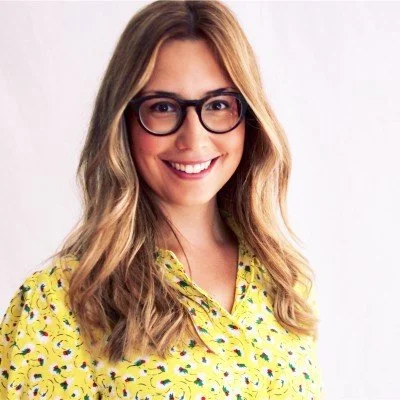 A woman with long, wavy blonde hair wearing glasses and a yellow blouse with a colorful pattern, smiling against a white background.