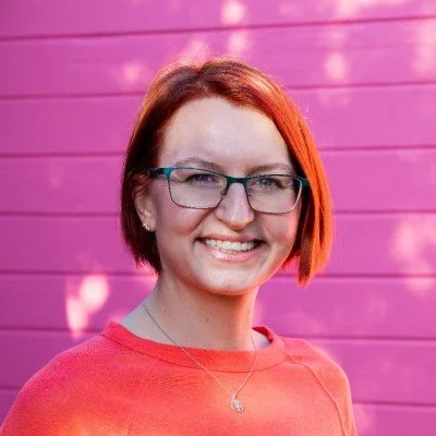 Young woman with red hair, glasses, and a bright smile standing in front of a pink wooden wall.