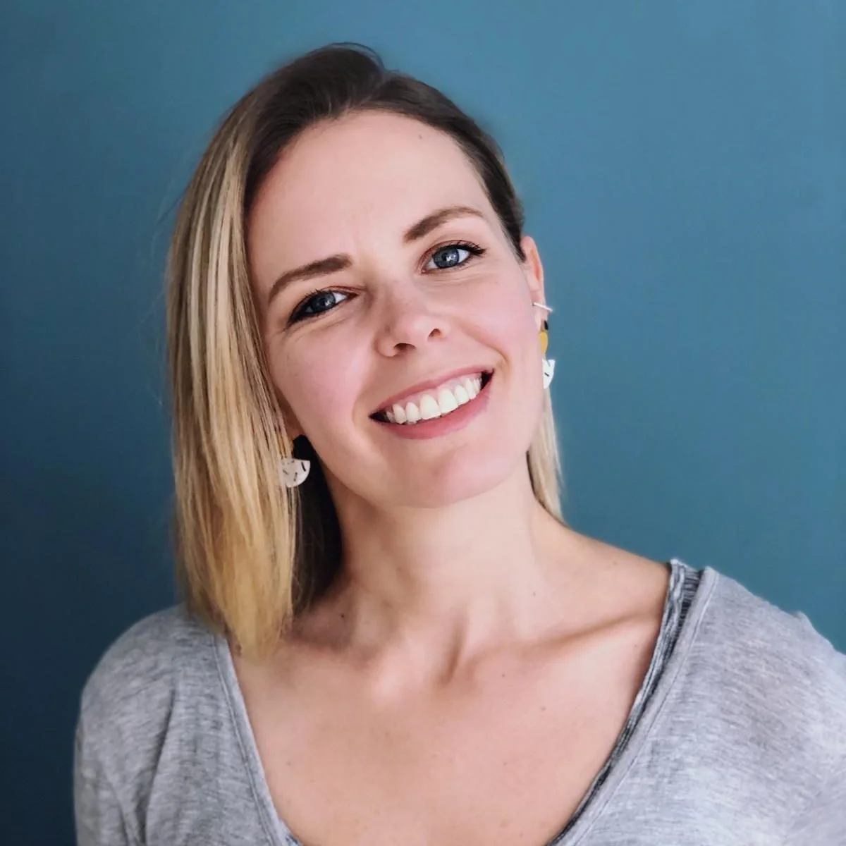 A woman with shoulder-length blonde hair, blue eyes, and a bright smile, wearing earrings and a light gray top, standing against a solid blue background.
