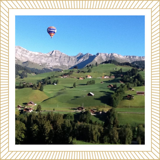 A hot air balloon floating over a lush green hillside with scattered houses, with mountains in the background and a clear blue sky.