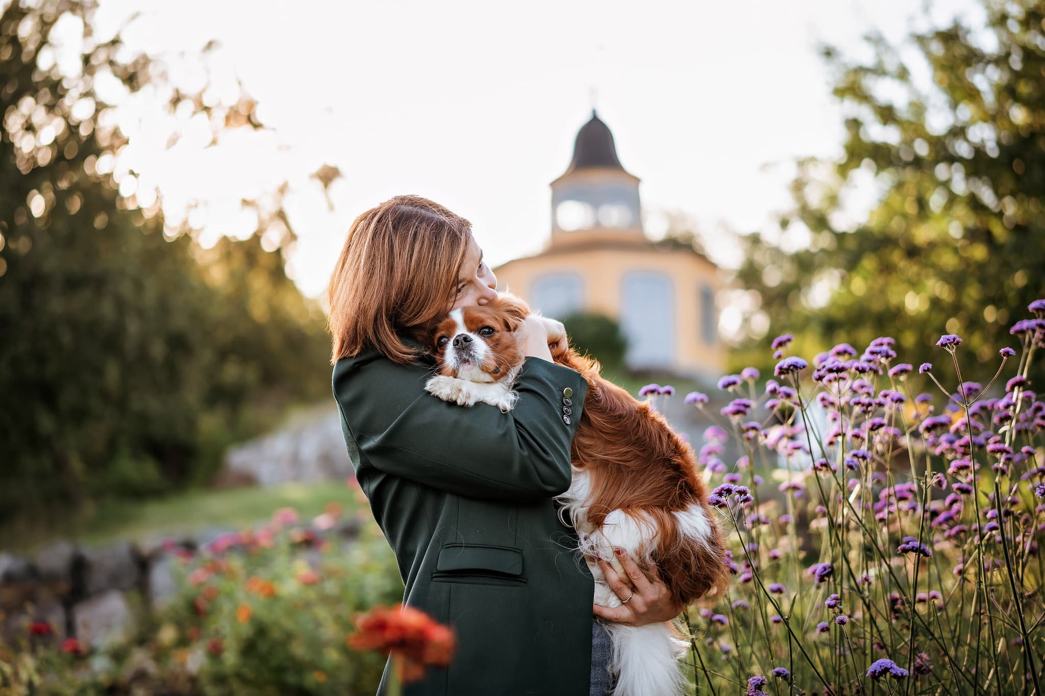 Woman holding a brown and white dog in a garden with colorful flowers, with a yellow building and trees in the background during sunset.