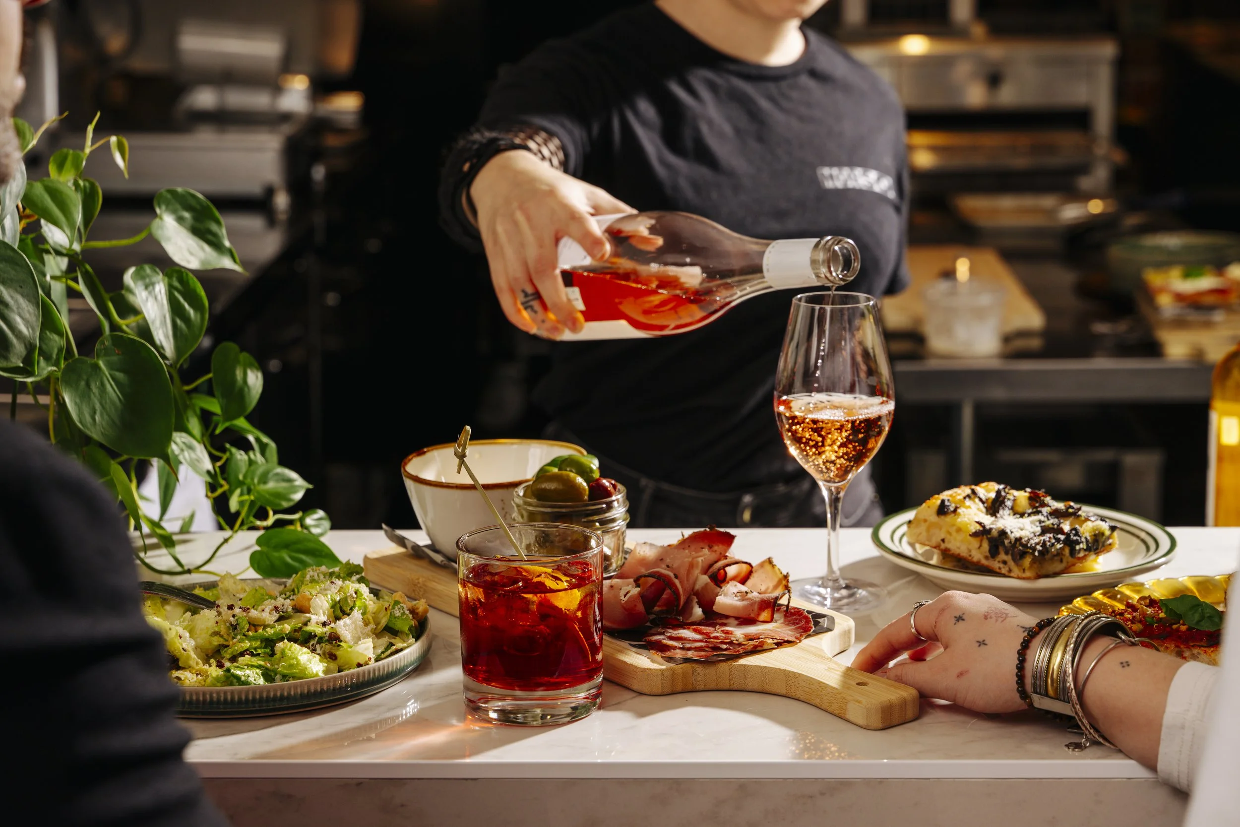 Une table avec de la nourriture et des boissons, un serveur verse du rosé dans un verre, une personne a la main posée sur la table, tout cela dans un cadre de cuisine ou de restaurant