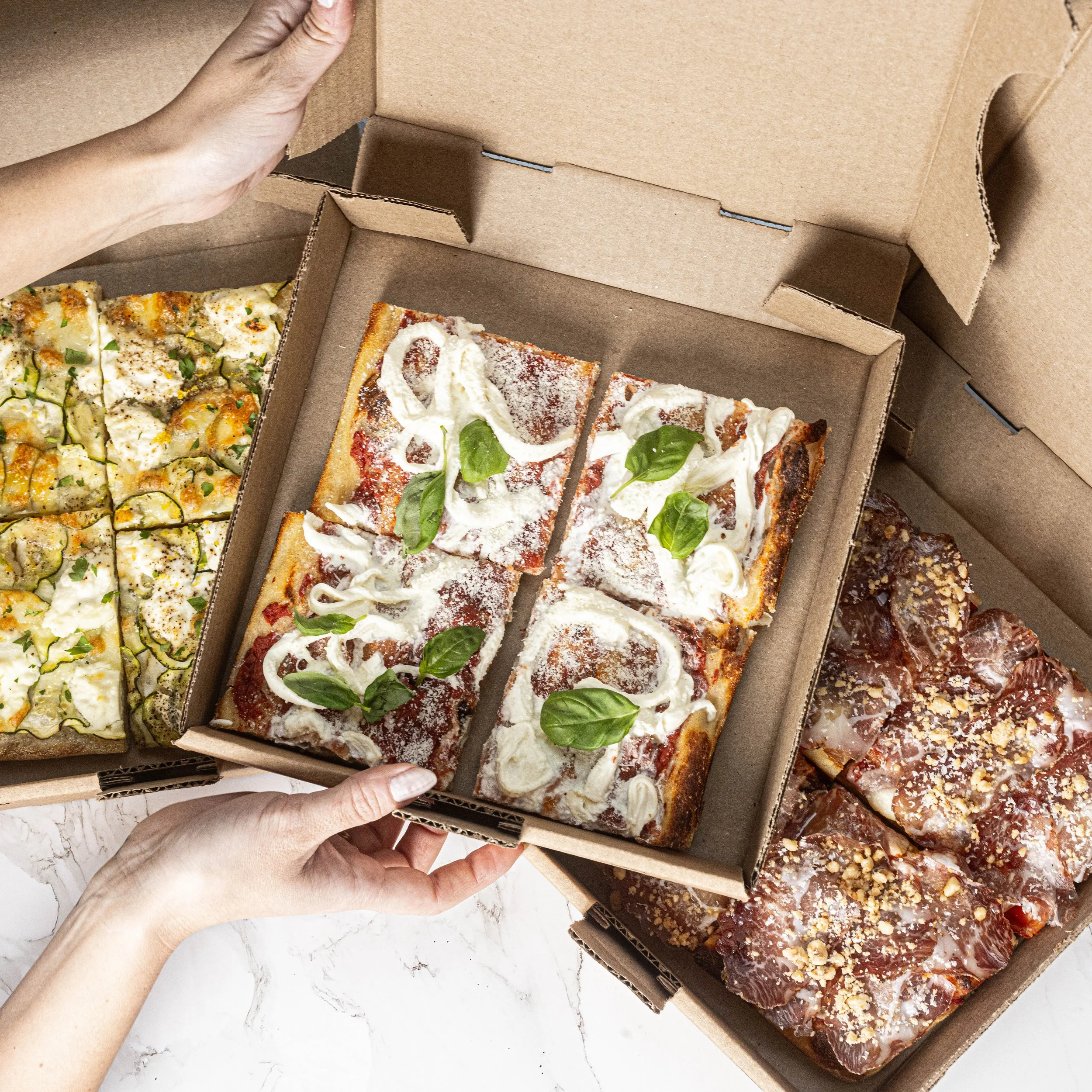 Boîtes de pizza avec différentes variétés, dont une pizza aux champignons, une pizza blanche avec fromage frais et basilic, et une pizza à la viande, vues d'en haut, sur une surface blanche.