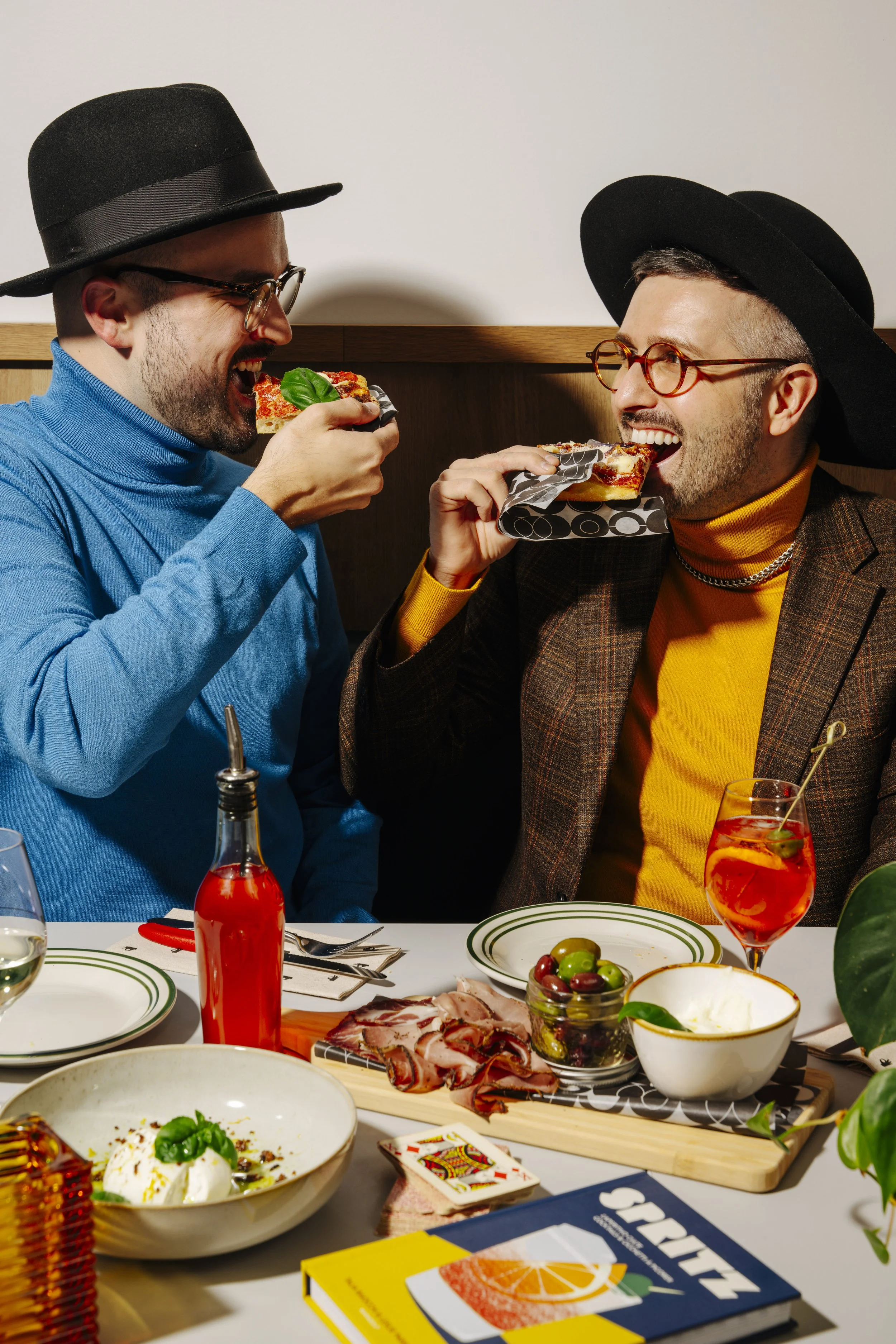 Deux hommes élégants avec des chapeaux noirs, portant des vêtements colorés, mangent des parts de pizza lors d'un repas convivial.