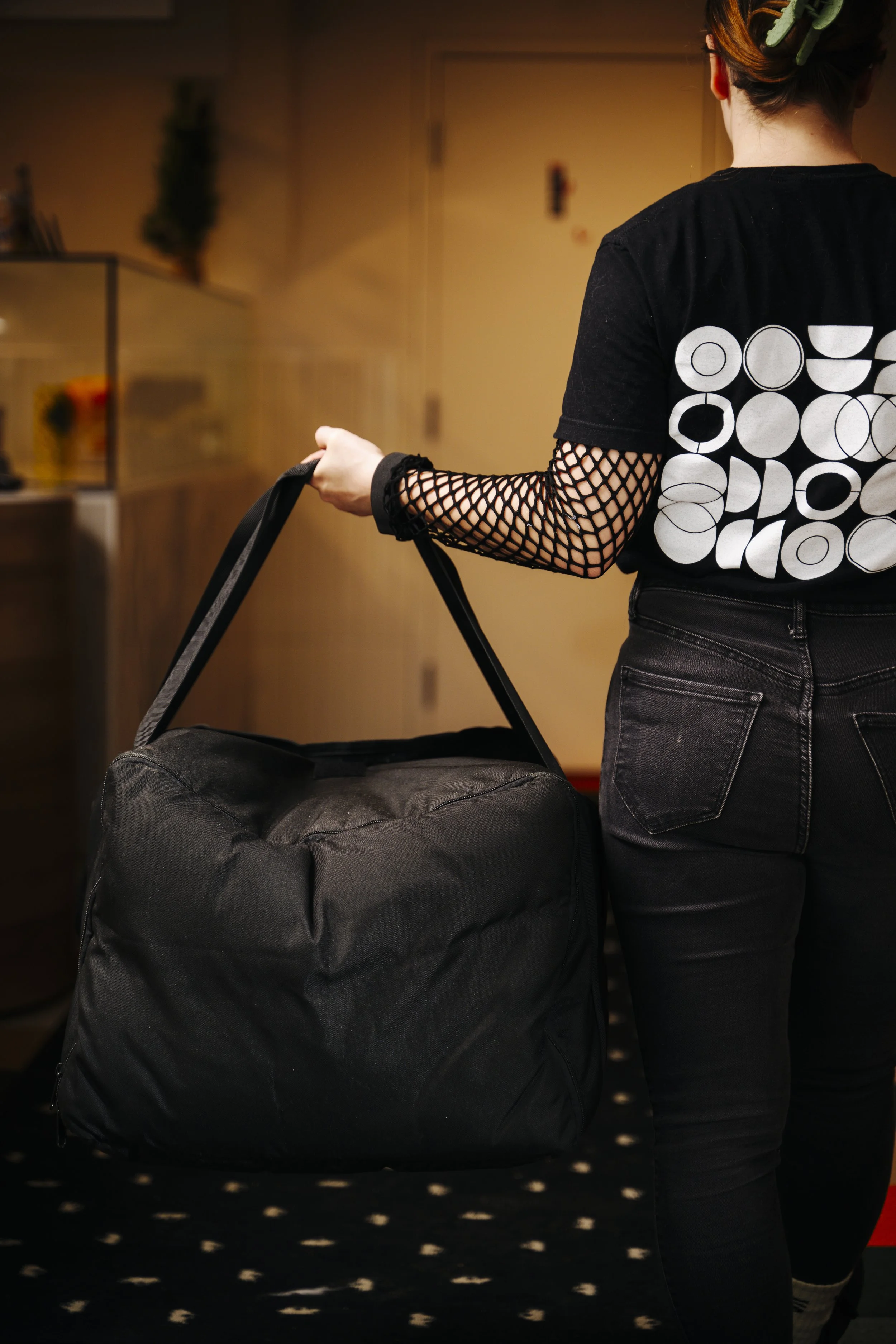 Femme portant un t-shirt noir à motifs géométriques, avec un filet en dentelle sur le bras, tenant un grand sac noir.