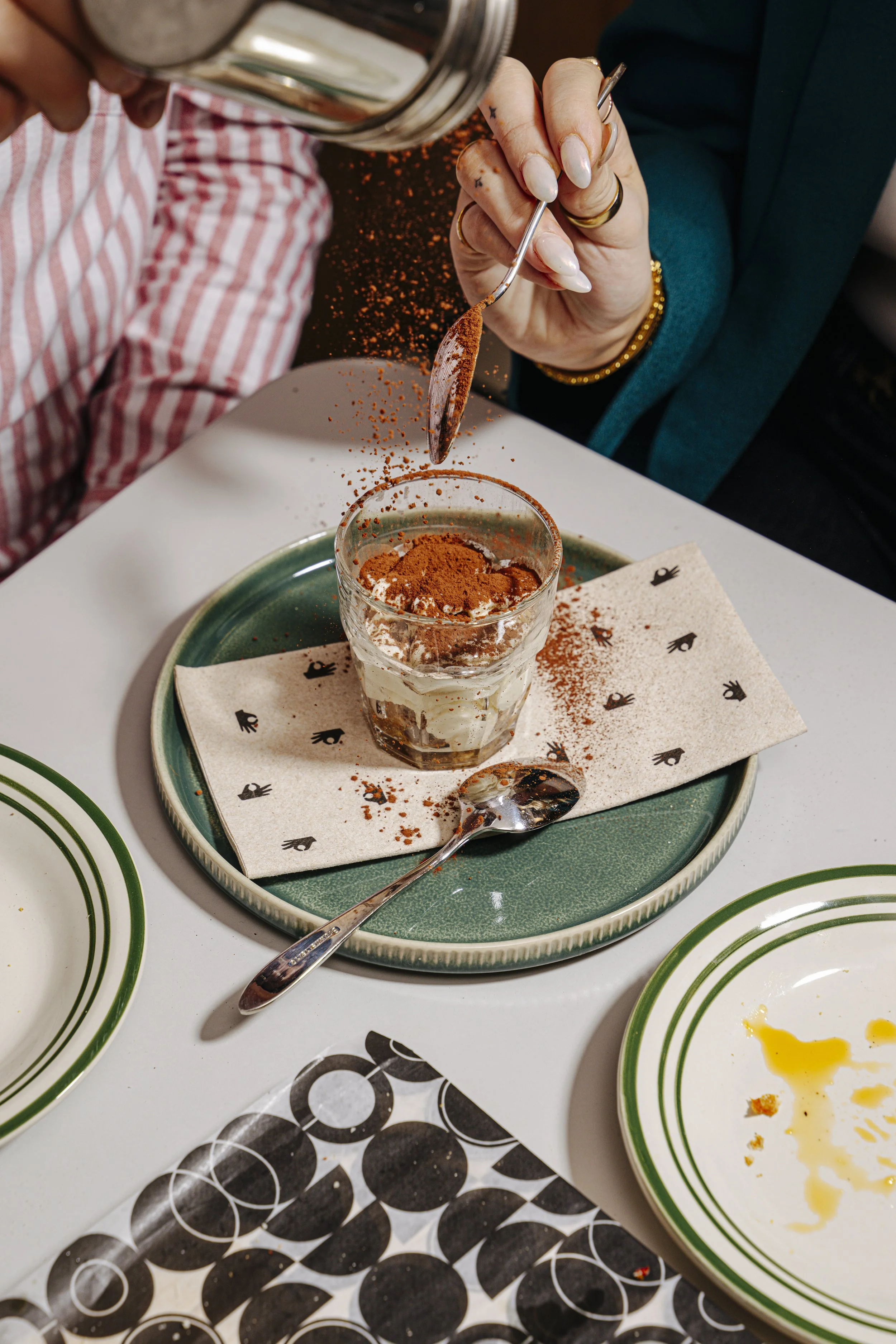 Une personne saupoudre de la poudre de cacao sur un dessert dans un verre, sur une assiette verte avec une nappe en papier à motifs. La table a aussi des assiettes blanches avec des bandes vertes, une cuillère en argent et un menu noir et blanc.