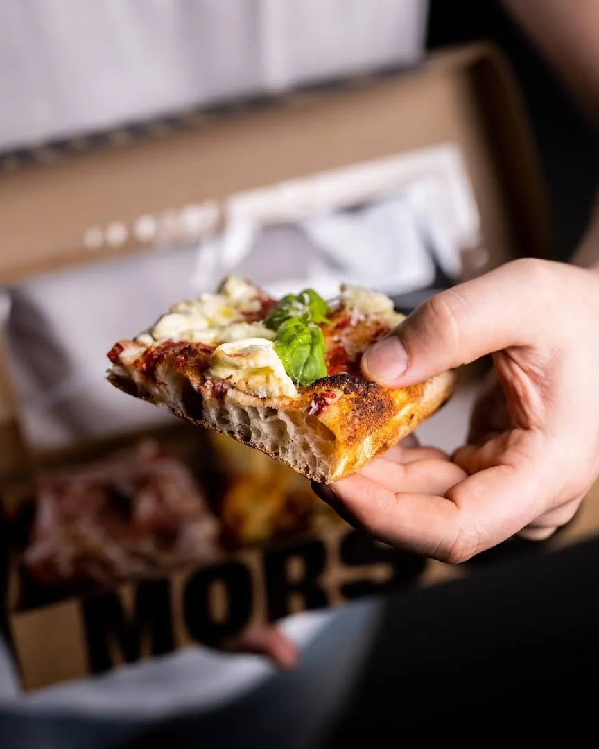 Une personne tenant une part de pizza avec du fromage, une feuille de basilic, de la sauce tomate et une croûte croustillante.