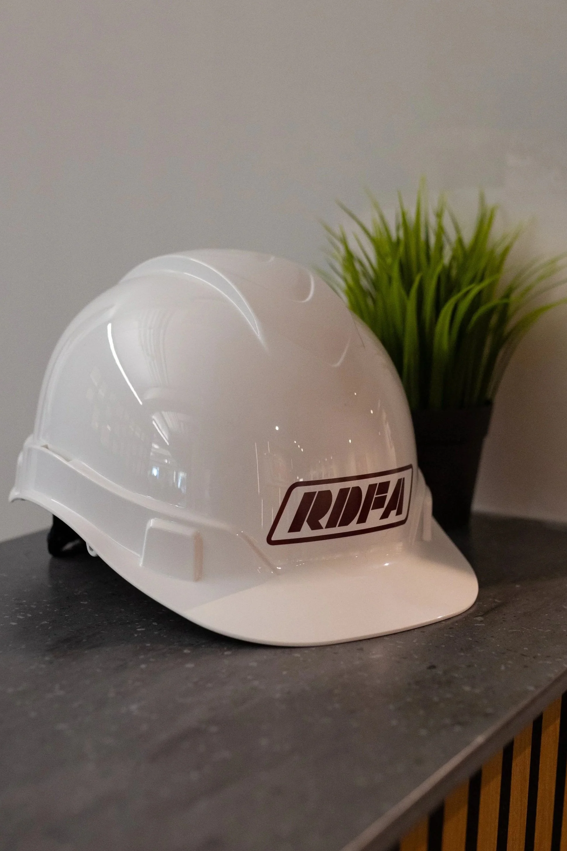 A white safety helmet with the logo 'RIDEA' placed on a black countertop, with a potted green plant in the background.