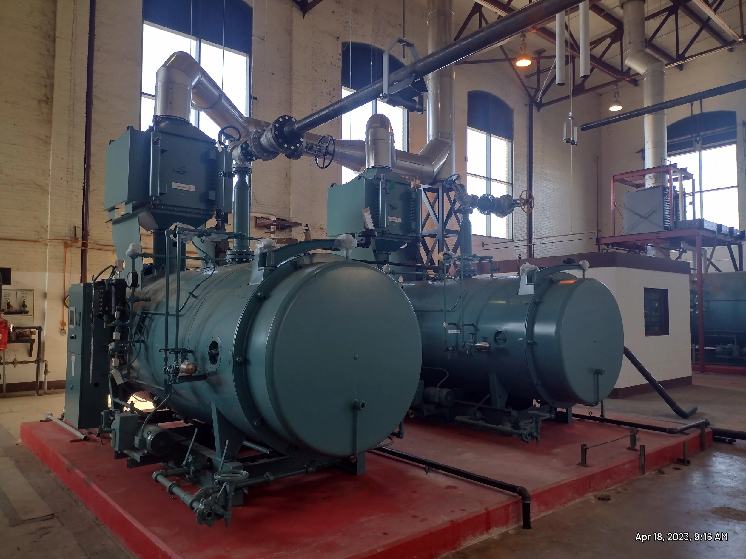 Industrial steam boiler system with large cylindrical tanks, pipes, valves, and controls inside a factory or plant, with high windows and high ceiling.