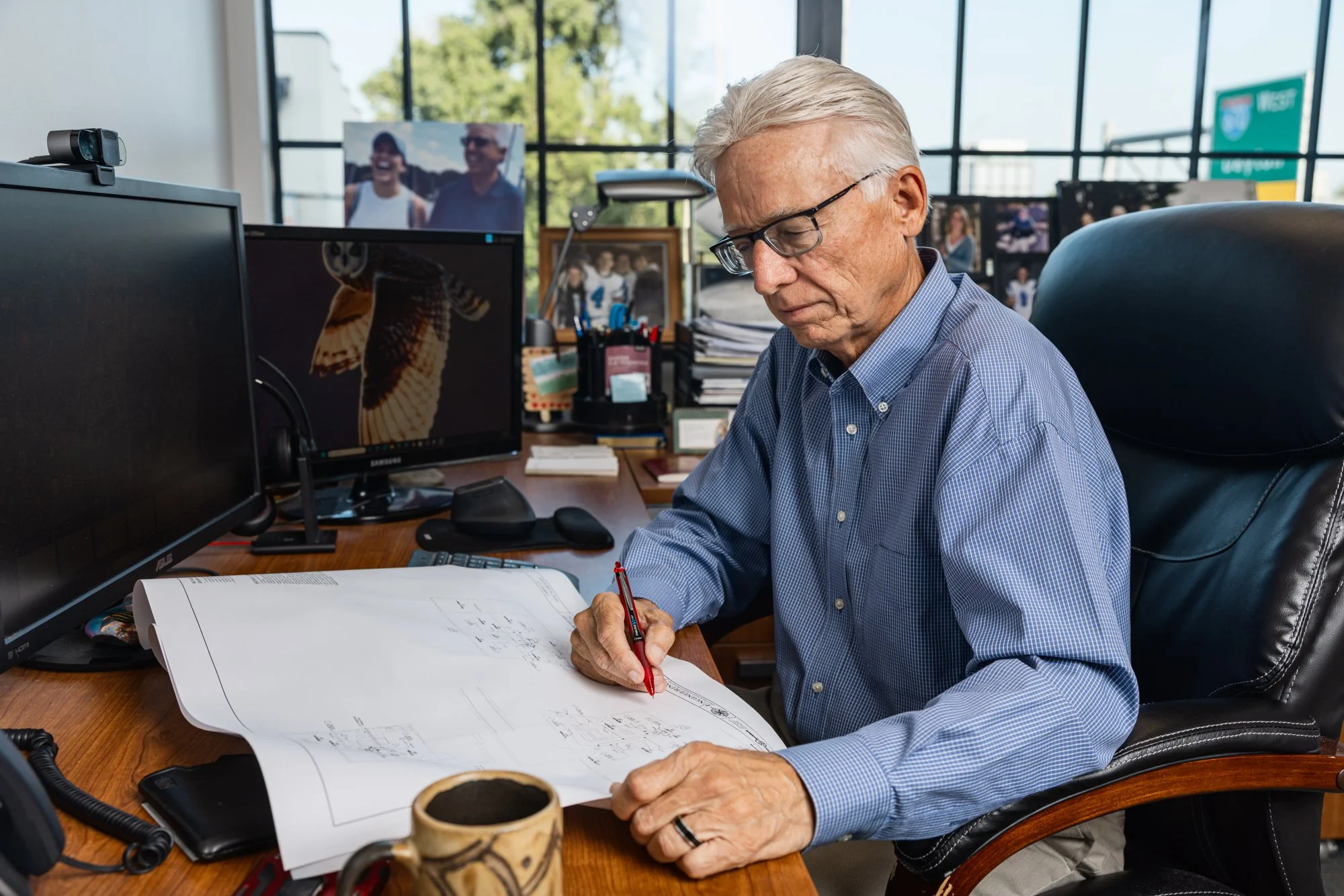 Greg Topp working at his desk, reviewing blueprints and taking notes with a red pen, surrounded by office equipment, multiple monitors, framed photos, and a coffee mug.