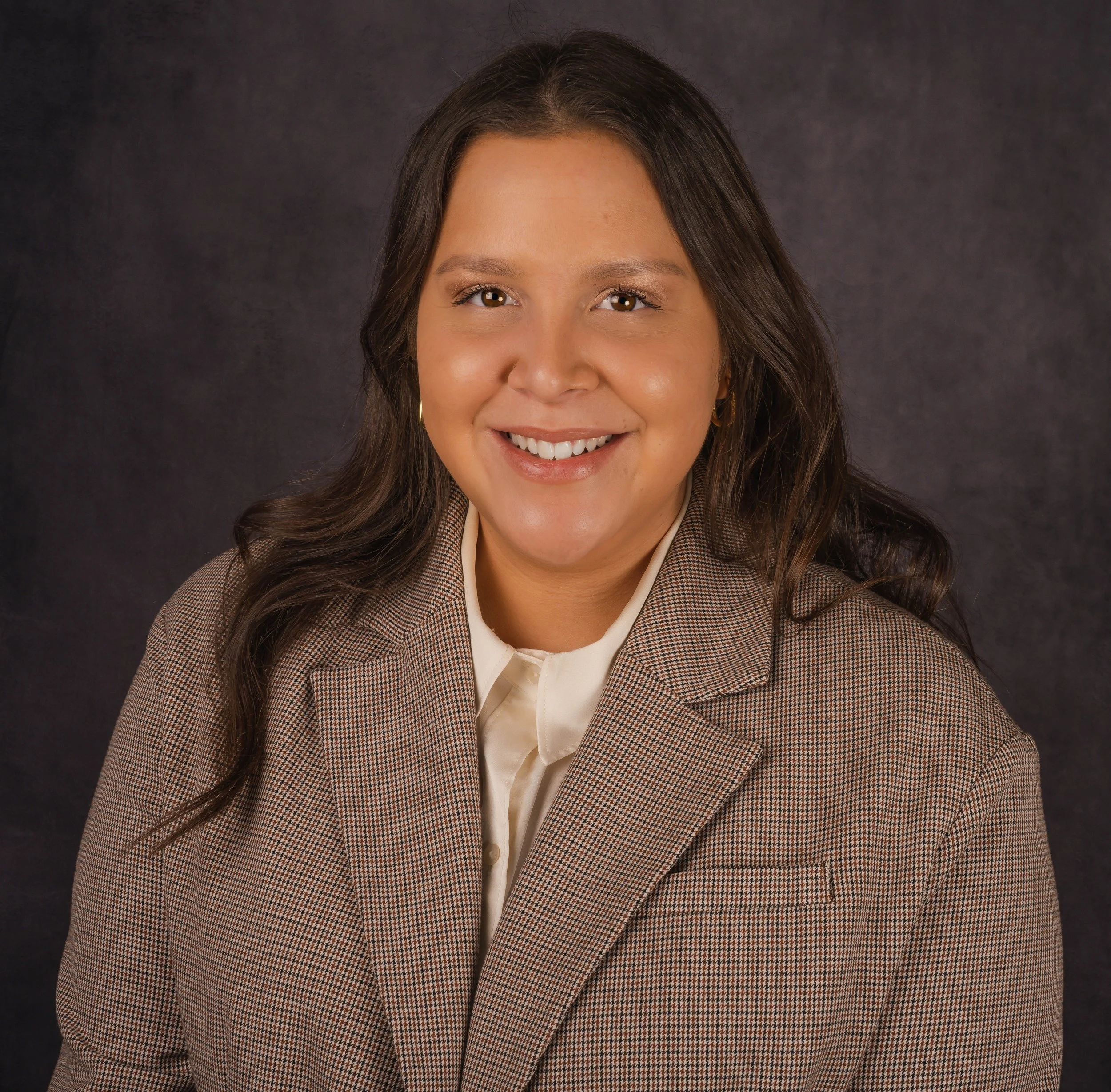 A woman with long brown hair smiling, wearing a brown coat, sitting indoors with a grey background.