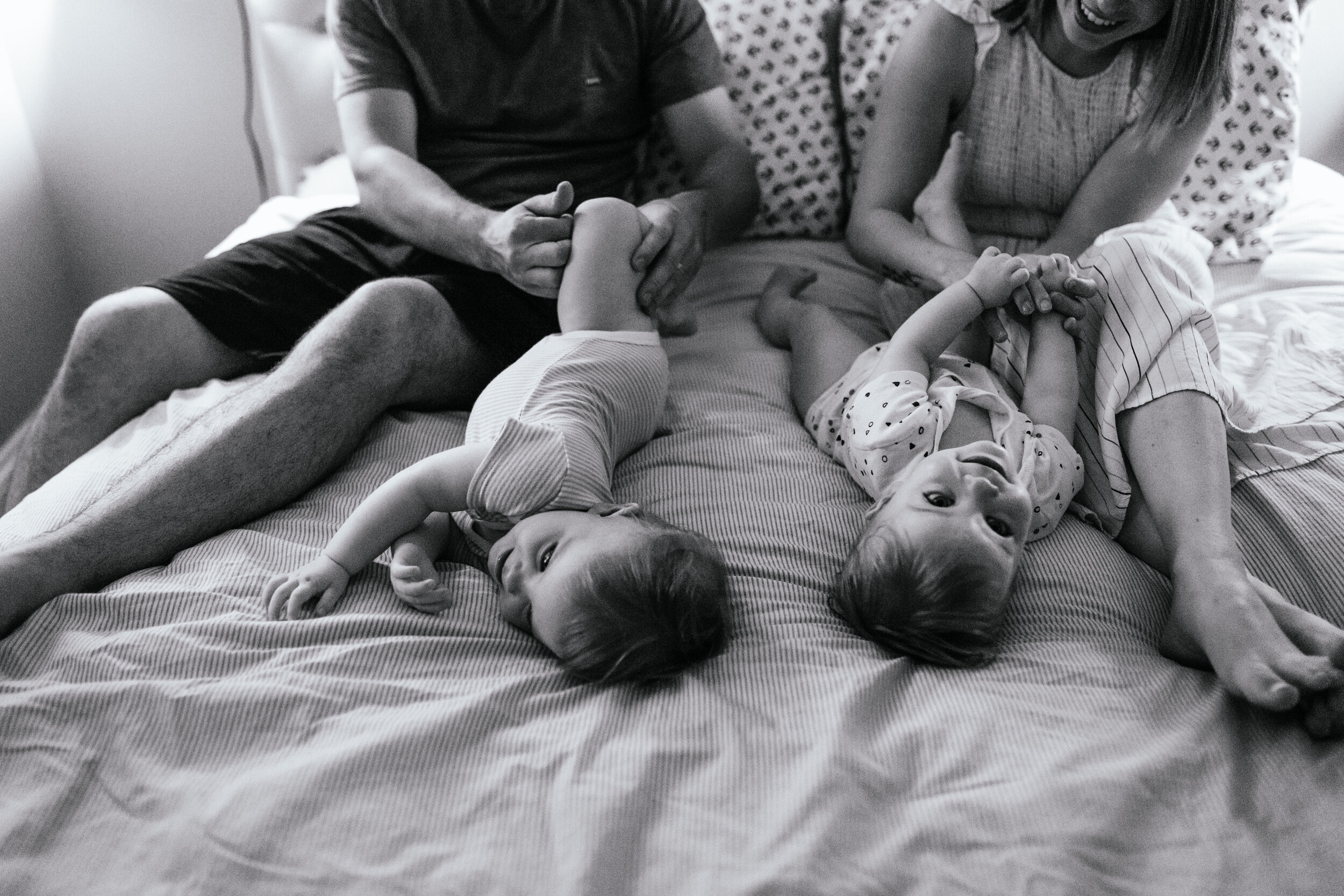 Black and white photo of a family on a bed, featuring two toddlers lying down and parents sitting nearby, engaged with the children.