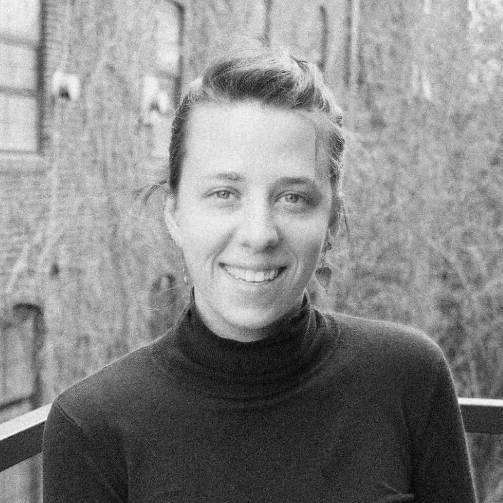 Black and white photo of a person smiling, with only the upper portion of their face visible, showing hair styled in a bun, eye glasses, and a tank top.