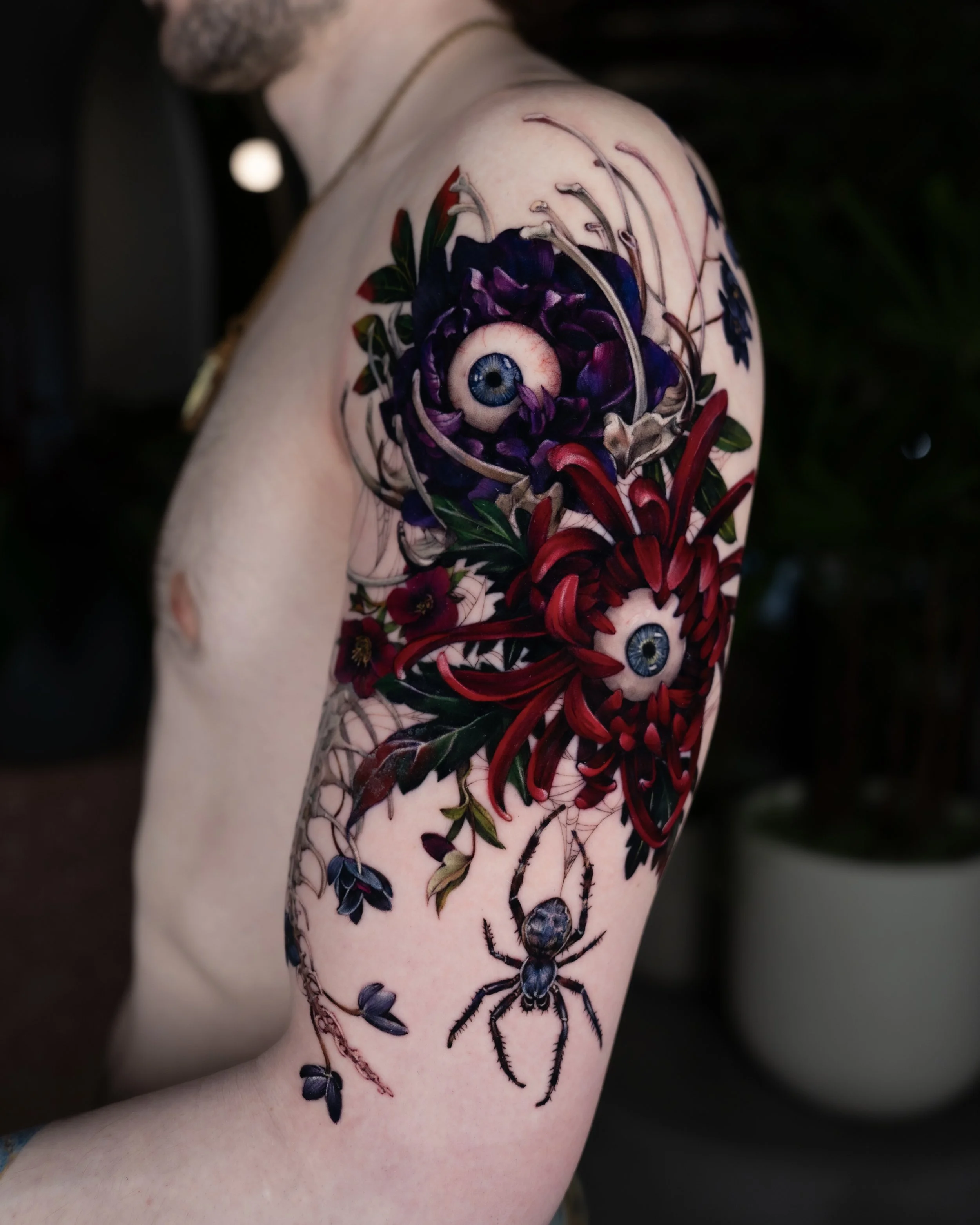 Tattoo of a large spider with multiple eyeballs on its back, covering the shoulder and upper arm area, with dark purple, black, and red colors, surrounded by green leaves and red flowers.