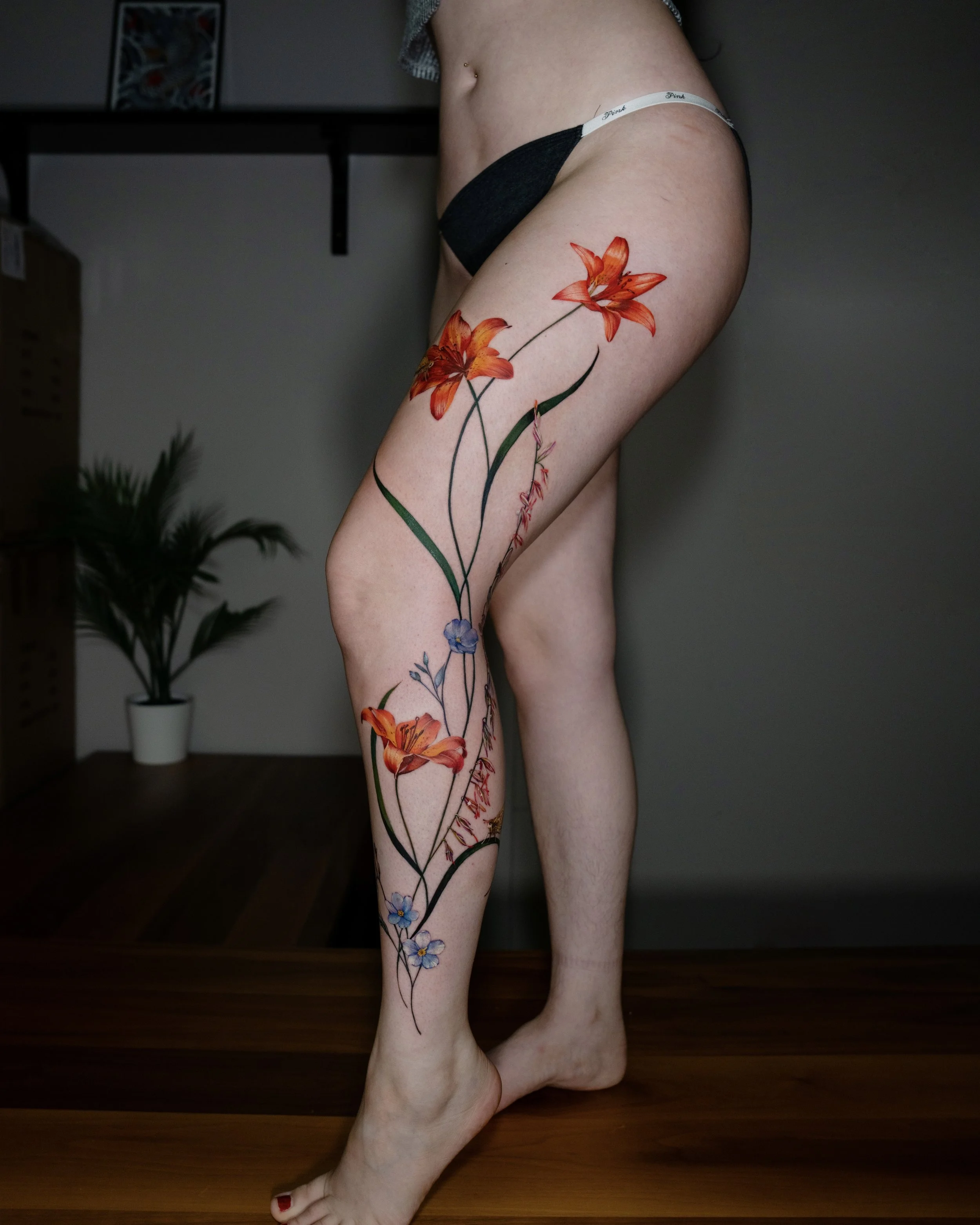 A person's leg with a detailed floral tattoo featuring orange, yellow, blue, and purple flowers extending from the upper thigh to the ankle.