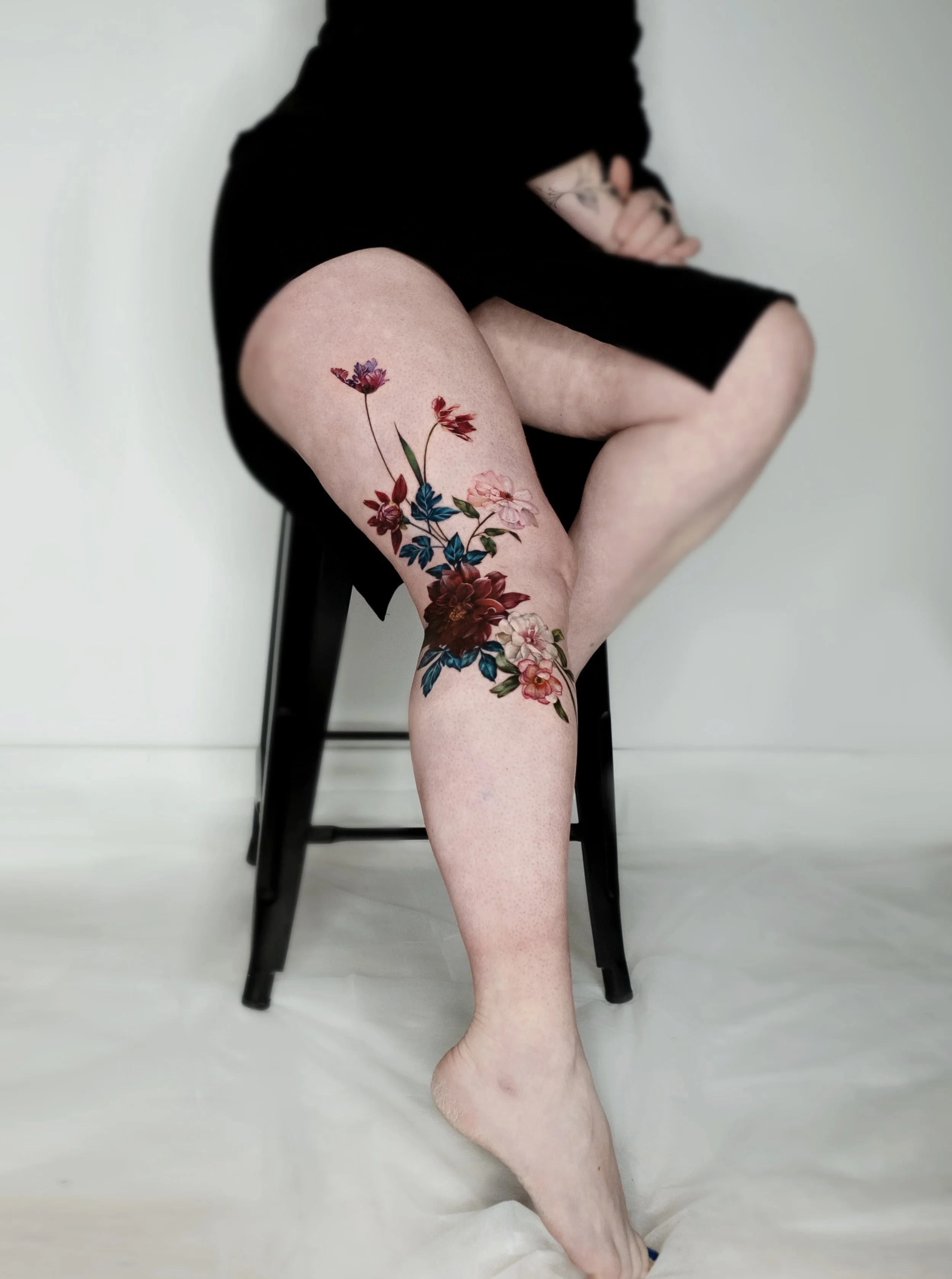 Person sitting on a black chair with one leg crossed over the other, showing a colorful floral tattoo on the thigh, wearing black clothing, and having their face covered, in a minimalistic setting with a plain background.