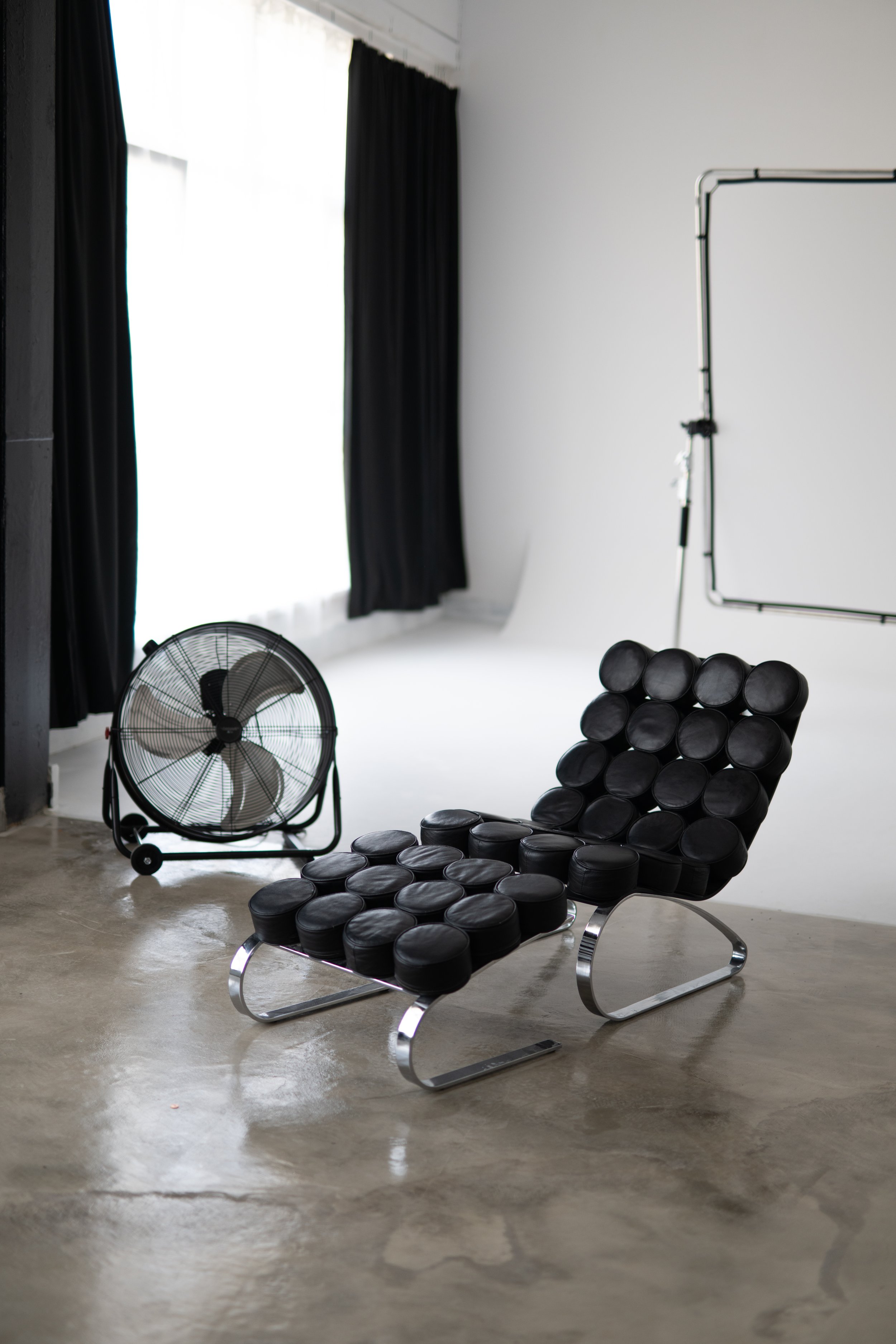 Modern black lounge chair with a rounded design made of multiple circular cushions and a chrome frame, positioned on a polished concrete floor. In the background, a fan on the floor, black curtains covering a large window, and a white wall with a bla