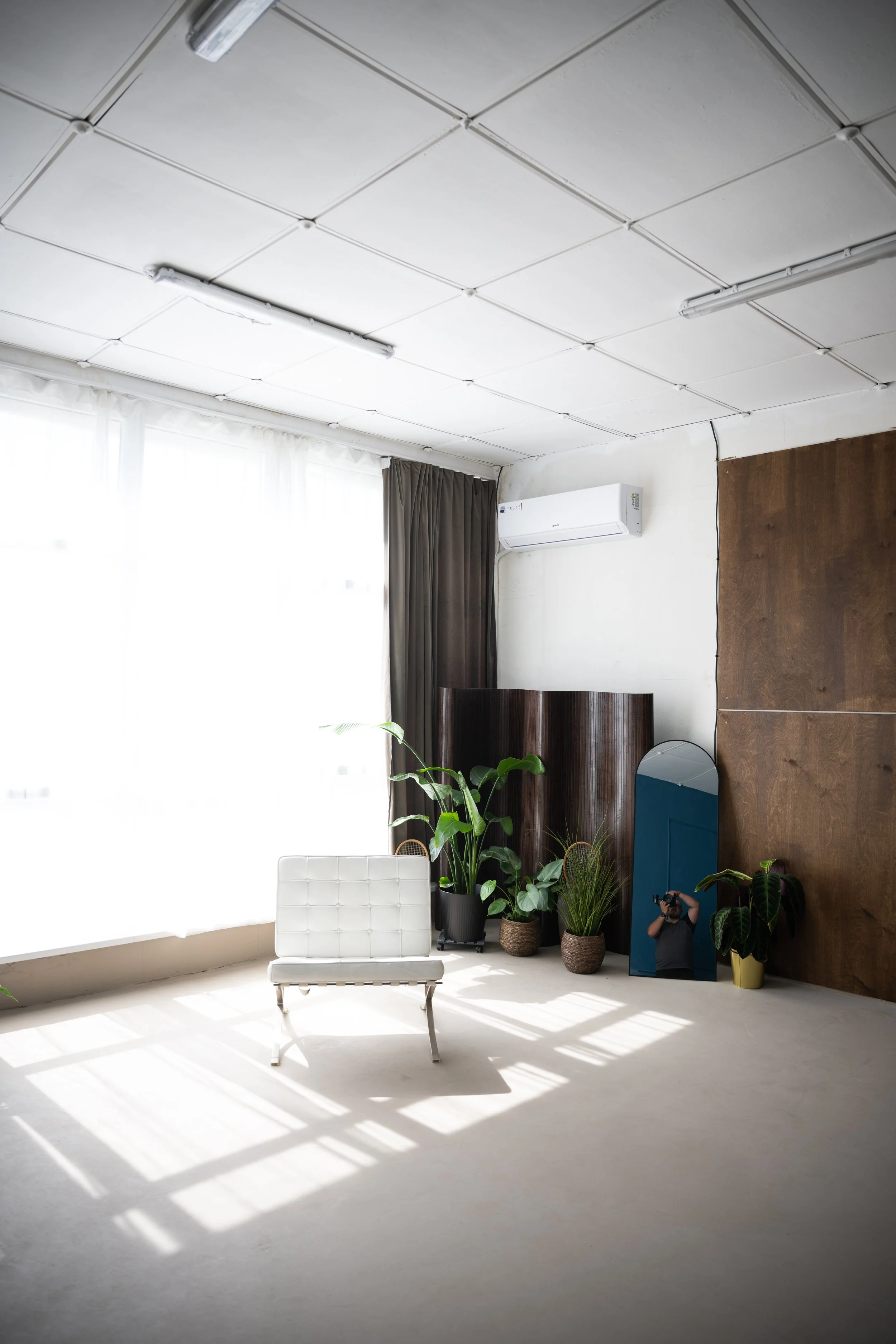 Bright room with natural sunlight, white floor, white ceiling with fluorescent lights, and a white chair. There are several potted plants near a large window with white curtains and dark drapes. A mirror and wall-mounted air conditioner are also visi