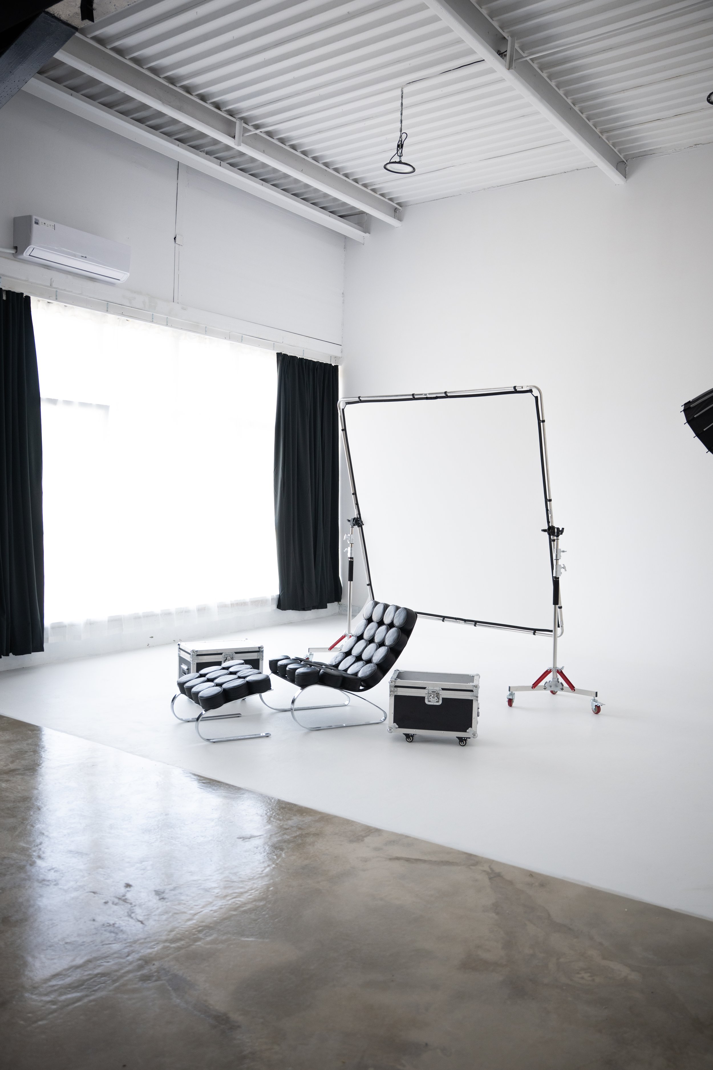 A photography studio with a white screened backdrop, black curtain, a modern black lounge chair with a matching footstool, lighting equipment on stands, a metal case, and a window with curtains in the background.