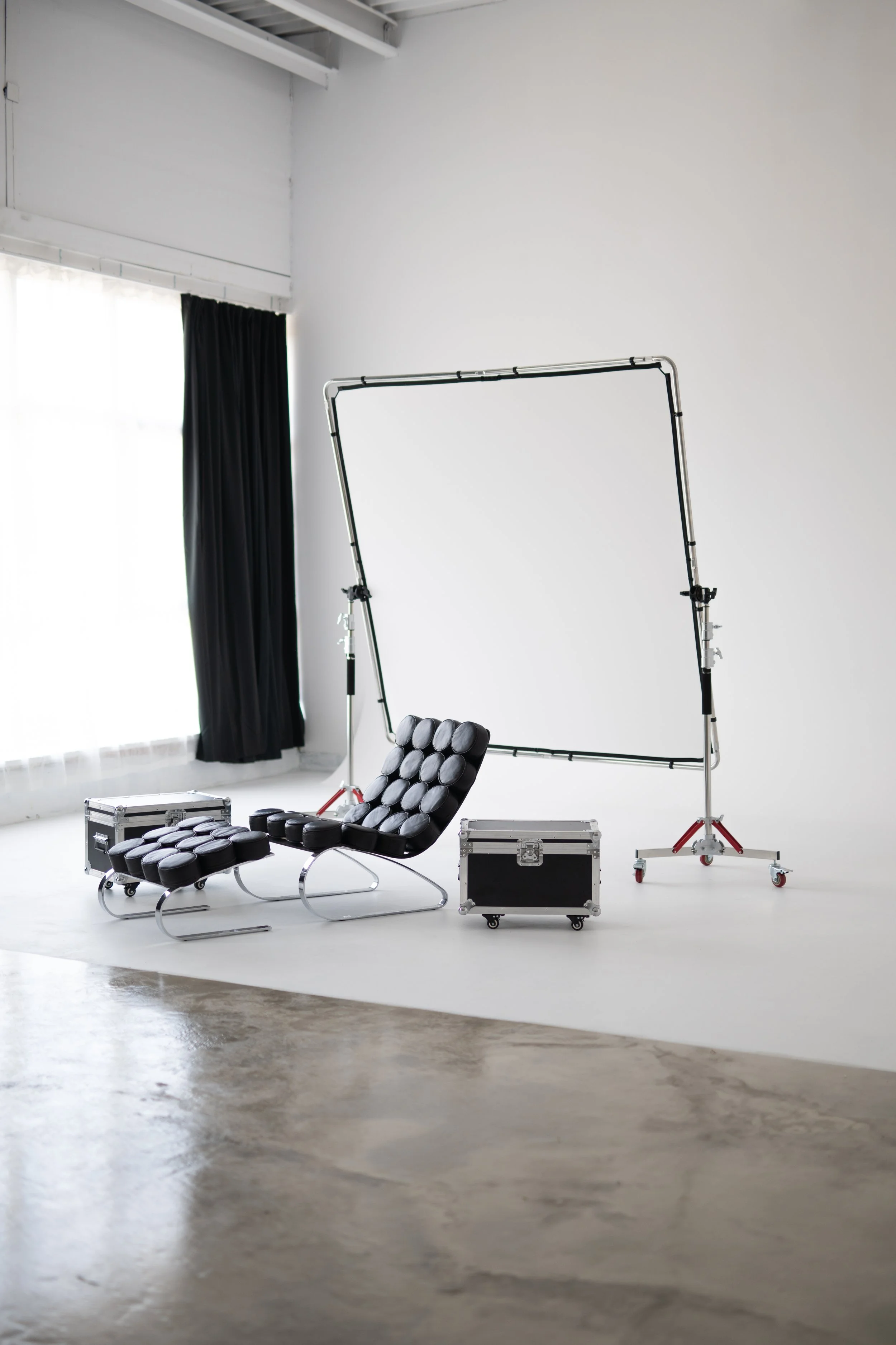 A photo of a photography studio with a white background, black curtains, and photography equipment including a black adjustable chair with circular padded cushions, a metal frame for lighting or backdrops, light stands, and equipment cases.