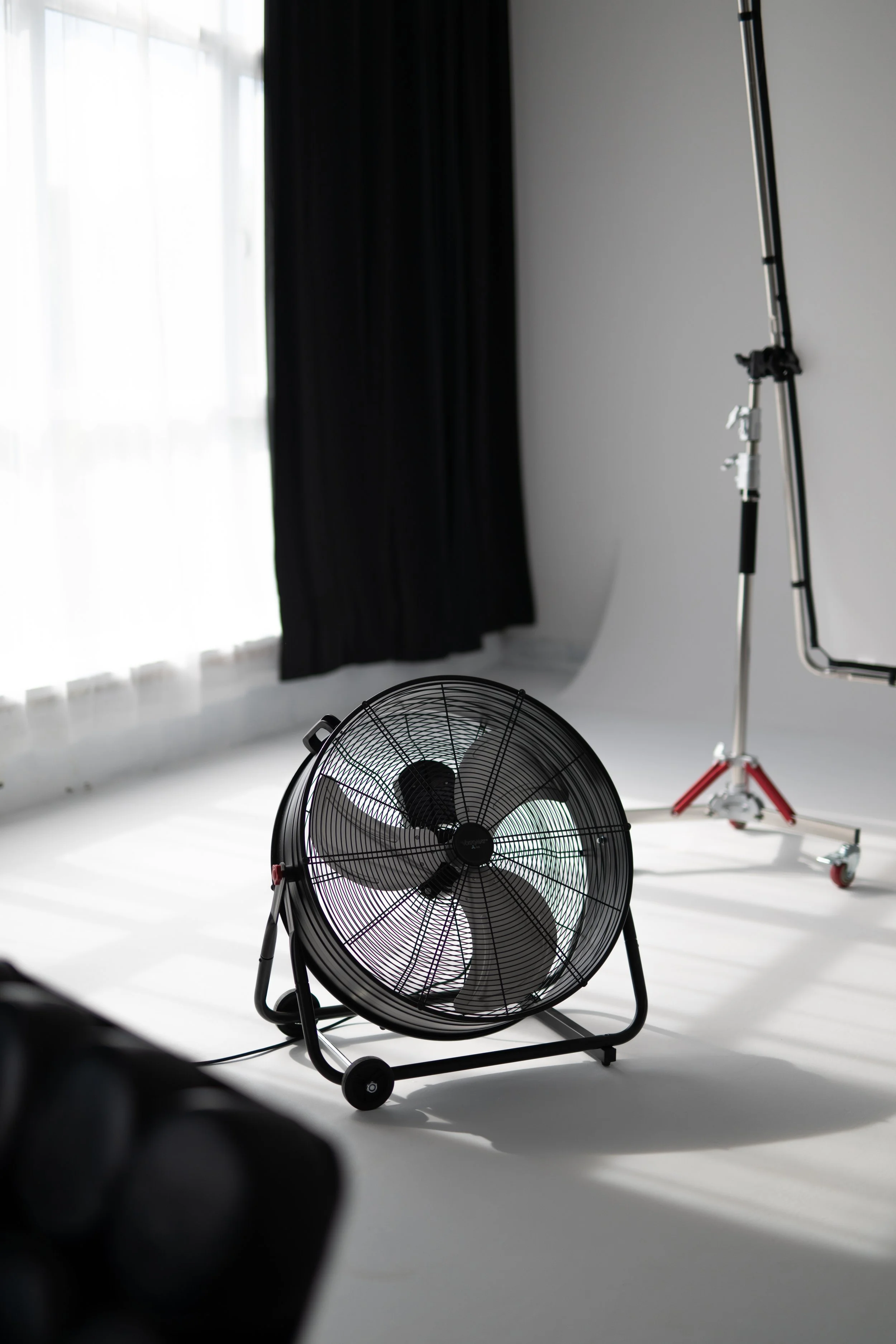 A photography studio scene with a black fan in the foreground, a white wall with black curtains on the left, and professional studio lighting and equipment in the background.