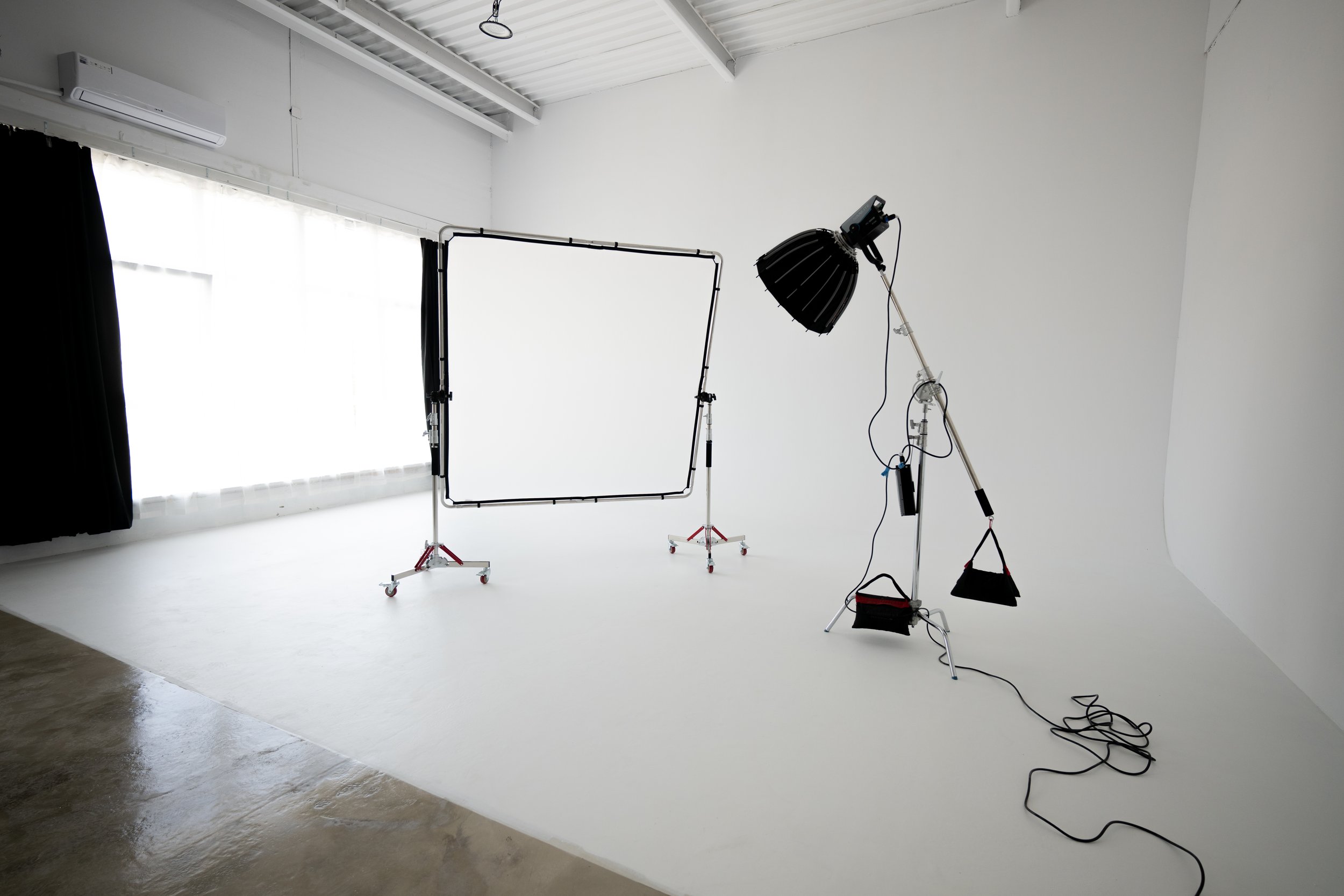 Photography studio with lighting equipment, a white backdrop, and a large window with black curtains in a bright, industrial-style room.
