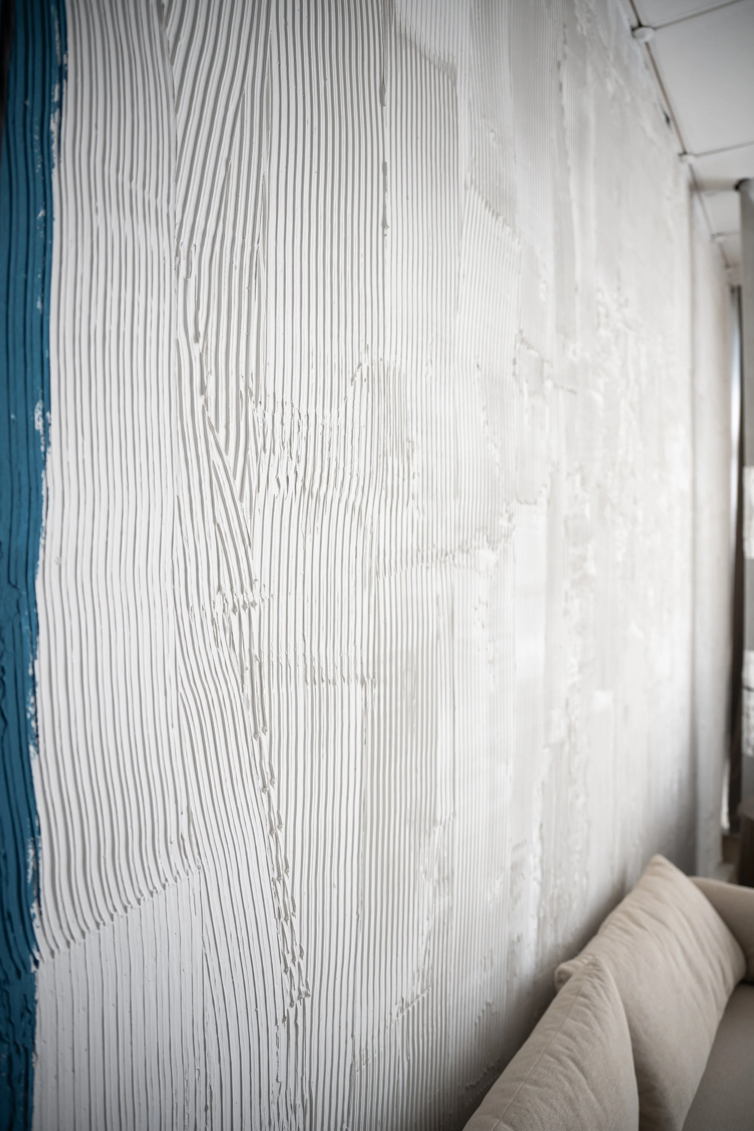 A wall partially covered with white textured plaster with vertical grooves, next to a beige sofa with cushions.