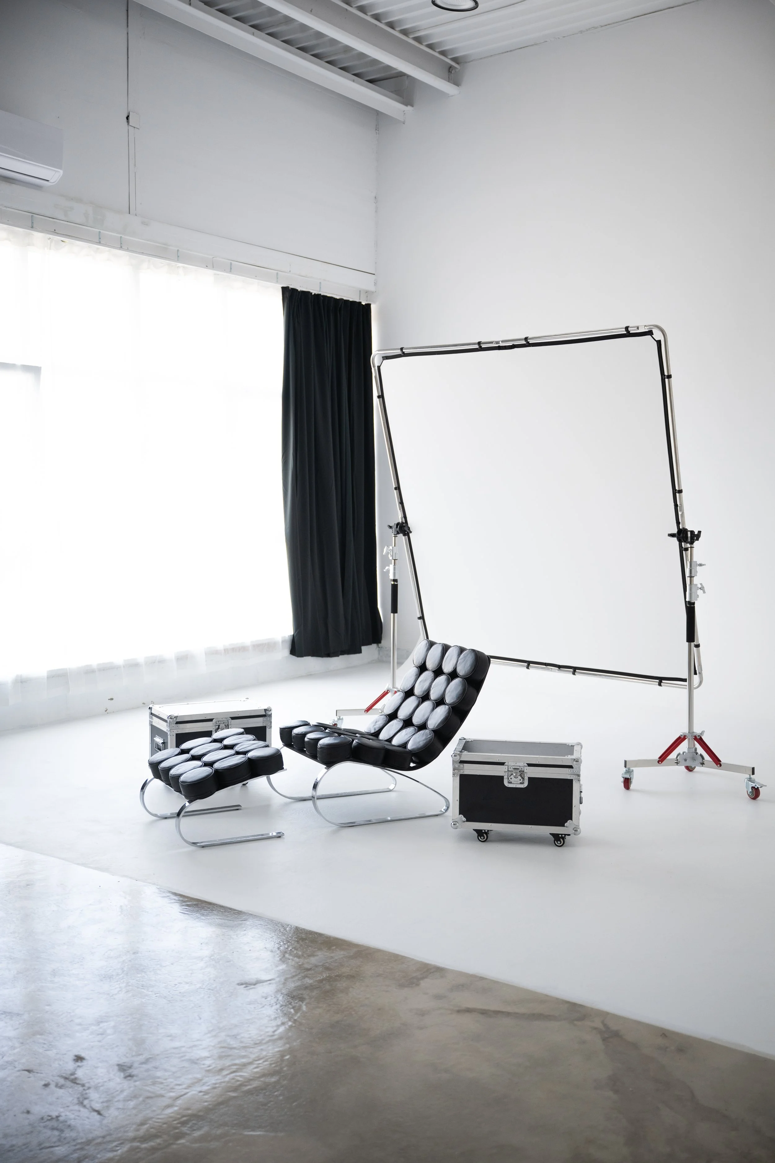 Photo of a photography studio with a black leather chair, multiple round cushions, a black case, and a large white backdrop frame. There is a window with sheer white curtains and a black blackout curtain. The room has a polished concrete floor and white walls.