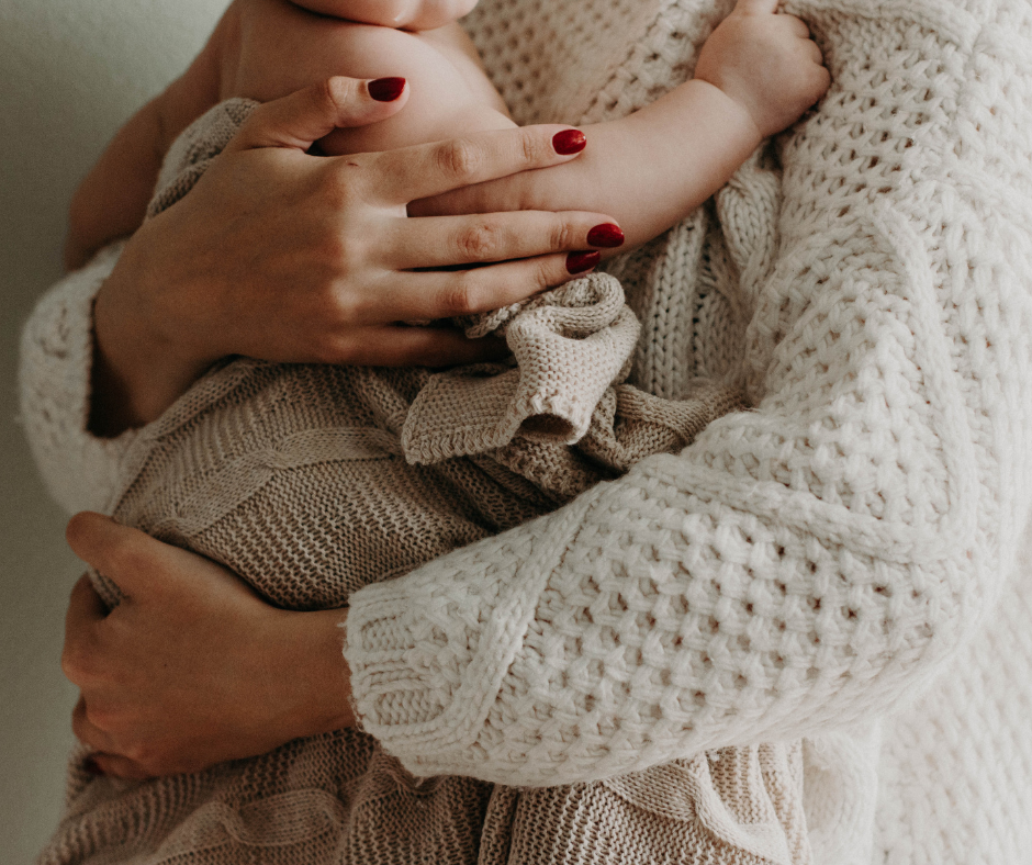 A woman with red nails and a cream cardigan holds a baby wrapped in a beige blanket