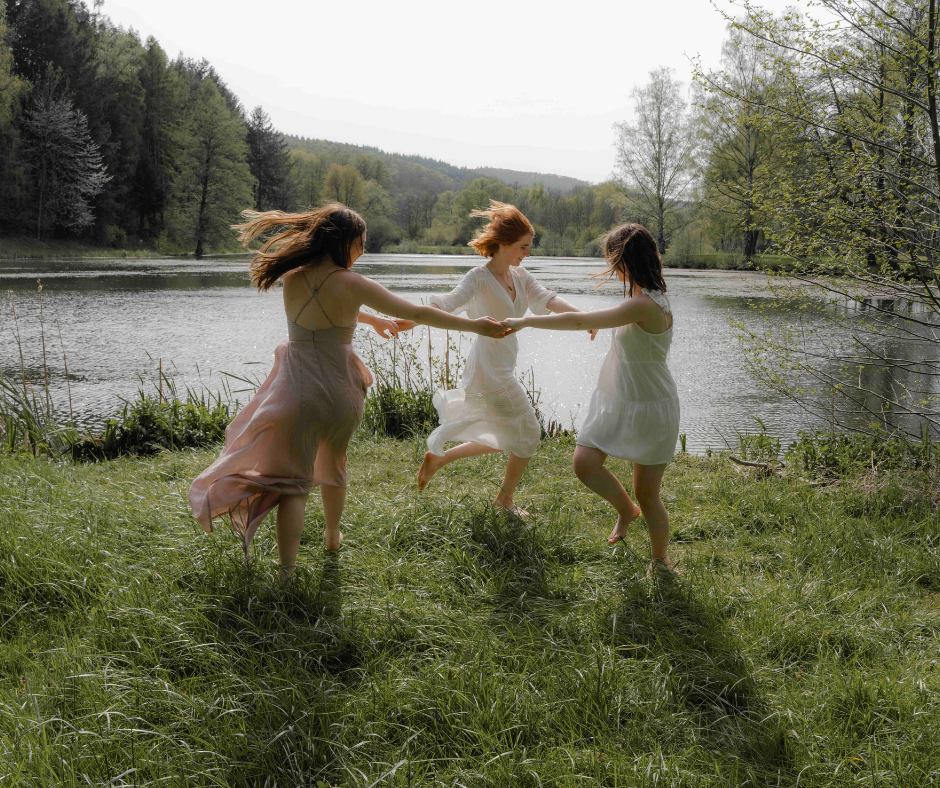 Three women in dresses holding hands and spinning in a circle by a lake