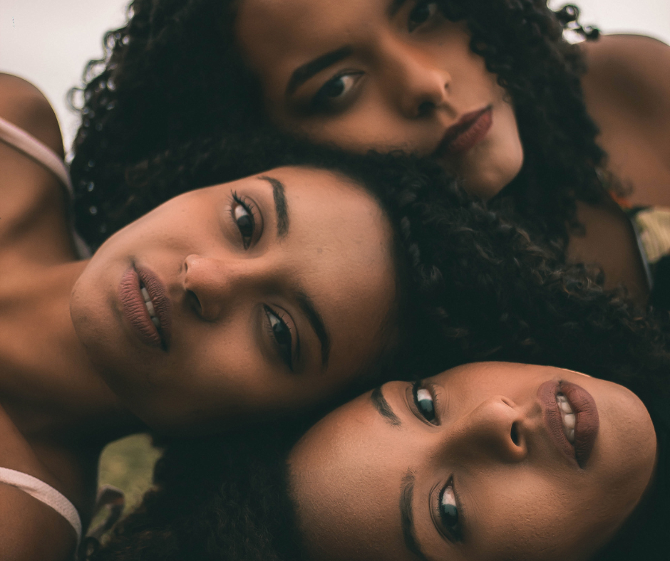 Three women lying closely together with their heads touching in a circle, all looking at the camera.