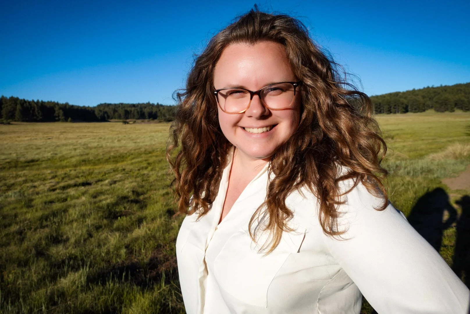 A woman with curly brown hair, glasses, and a white shirt smiling outdoors in a green field with trees and a blue sky in the background.