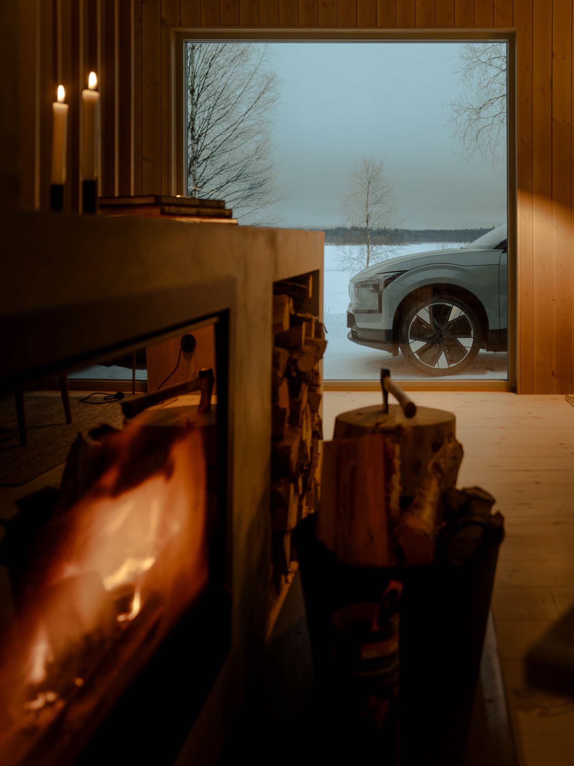 Indoor scene with a wood stove, fire visible through glass, logs in a large container, and a large glass window showing snow-covered landscape and a parked car outside.
